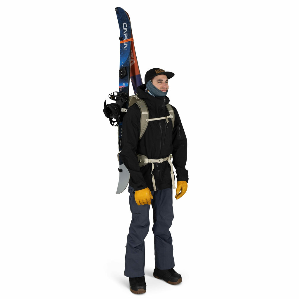 The image features a person dressed for outdoor adventure or possibly winter sports. They are wearing a black jacket, dark blue pants, and sturdy black boots. A pair of skis is strapped vertically to a light-colored backpack, suggesting preparedness for s