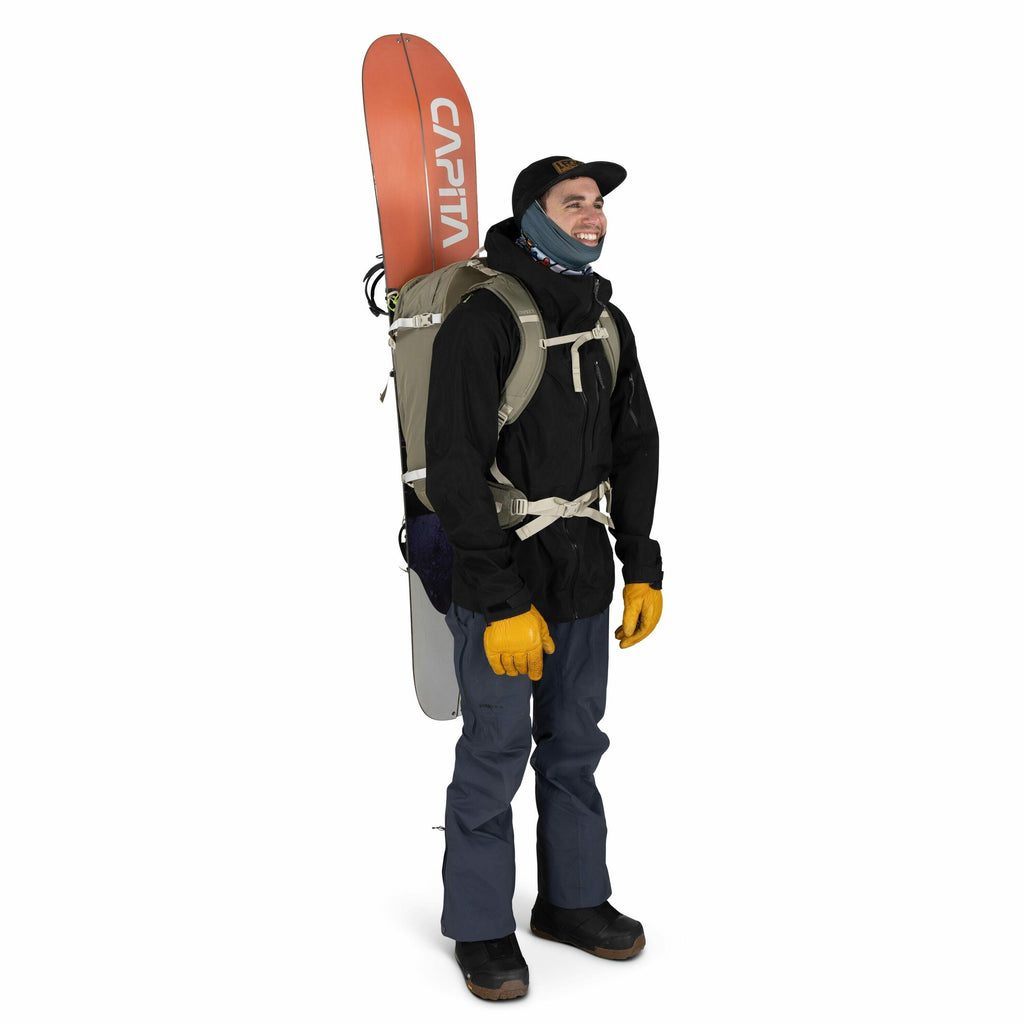 The image depicts a person standing and dressed for snowy outdoor conditions, possibly for snowboarding or skiing. They are equipped with a snowboard, branded with "CAPiTA," which is secured to a backpack. The person is wearing a black jacket, blue pants,