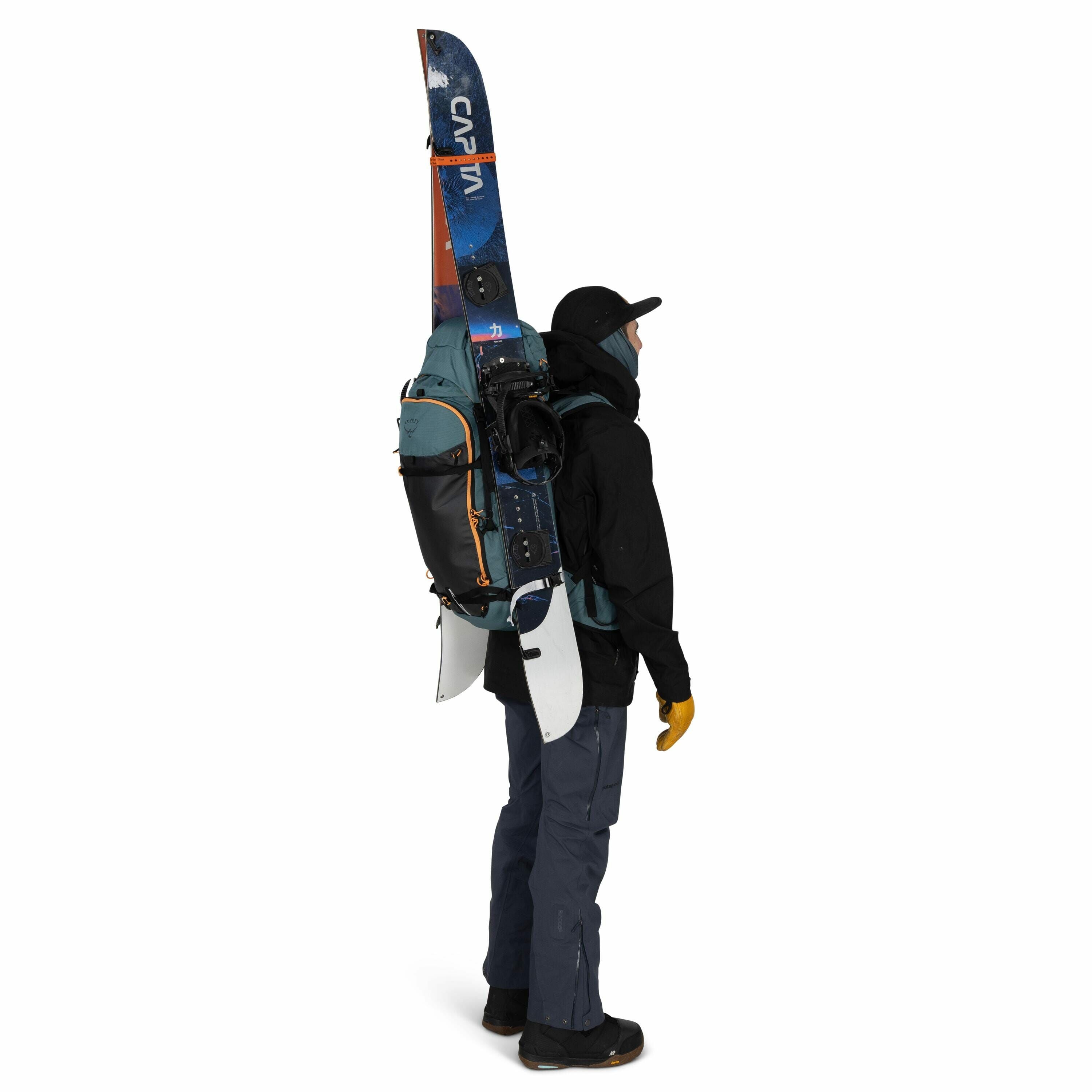 The image depicts a person standing in profile, fully equipped for snowboarding or skiing. They are wearing a dark jacket, dark pants, and black boots, with a black cap and yellow gloves for warmth. A snowboard is attached vertically to their large backpa