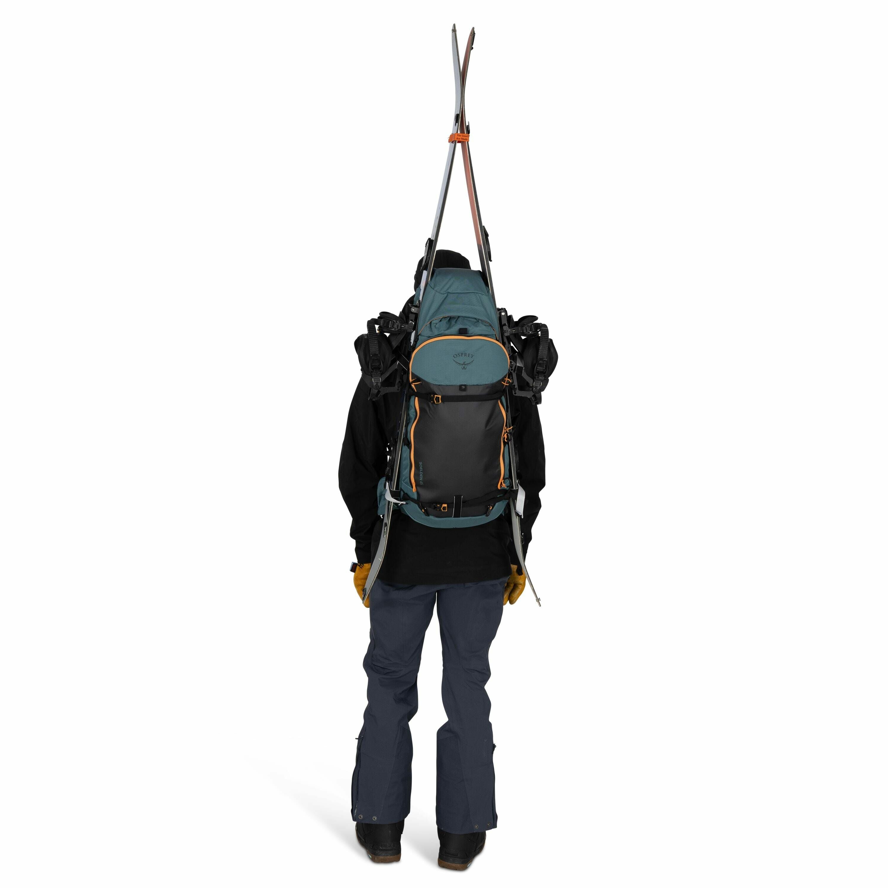 The image shows a person viewed from the back, who is fully equipped for a ski trip. They are wearing a large backpack, which has skis attached vertically on the back. The backpack is blue and gray with orange accents and appears to be made for outdoor ac