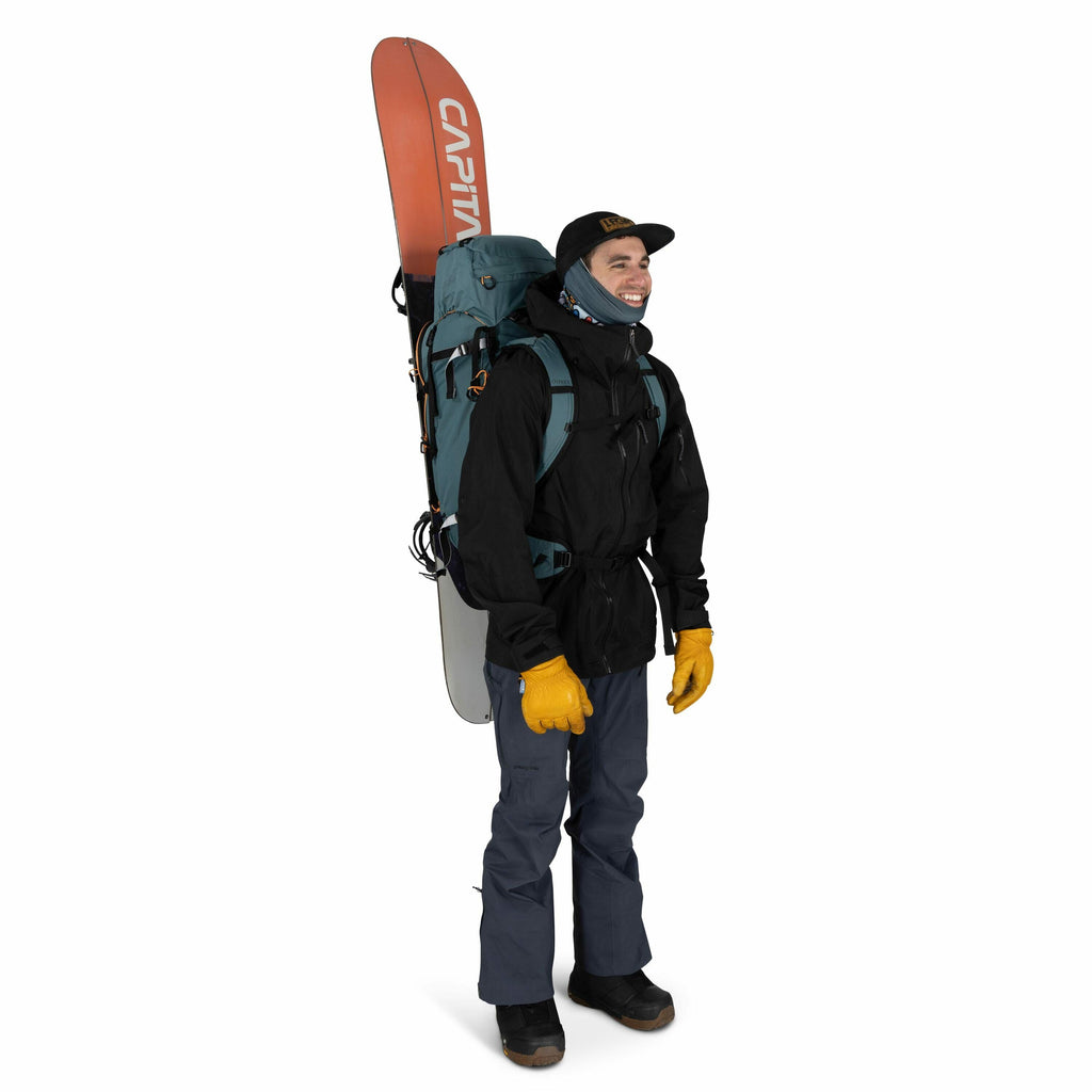 The image shows a person dressed in outdoor gear, likely prepared for a snowboarding or mountain adventure. They are wearing a black jacket, blue pants, and sturdy black boots, complemented by bright yellow gloves for protection against the cold. A blue b