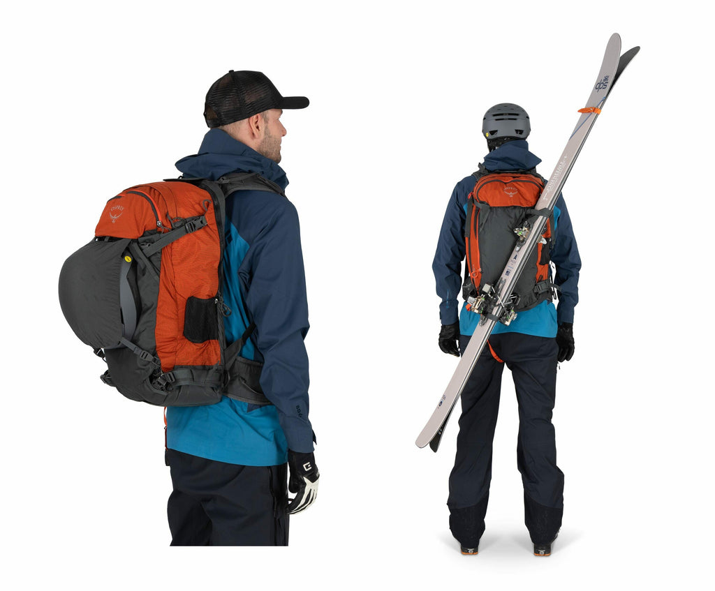 The image shows a person dressed in outdoor gear suitable for skiing or snowboarding. They are wearing a blue jacket over a teal layer, dark pants, and gloves. A large orange and grey backpack is strapped to their back, equipped with a helmet attached to