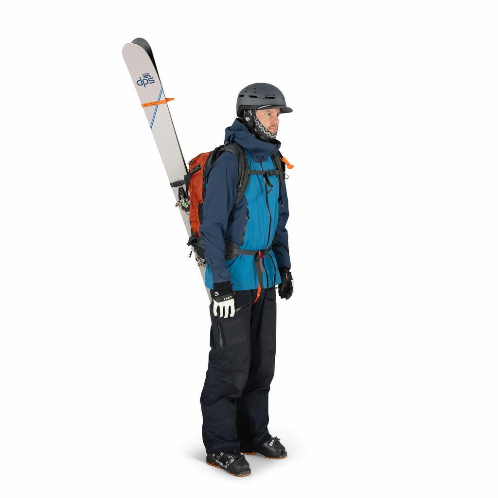 The image shows a person dressed in ski gear, ready for a day on the slopes. The individual is wearing a blue jacket over a darker blue base layer, black ski pants, and black ski gloves. They are carrying a pair of skis, which are mounted vertically on a
