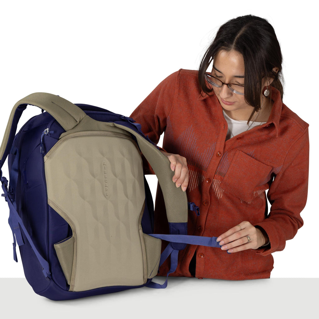 A person wearing an orange-red button-up shirt adjusts the blue strap of a large backpack. The backpack is navy with a tan, molded back panel that has the word "LIFTCAPE" embossed vertically. The person holds one shoulder strap with one hand and pulls the