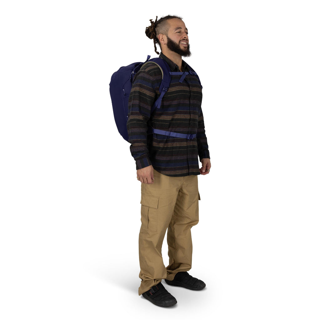 A person stands in a three-quarter pose wearing outdoor-style clothing and a backpack. They have on a dark striped button-up shirt, tan cargo pants, and black shoes. The backpack is navy blue with padded straps and a waist belt clipped at the front. The b