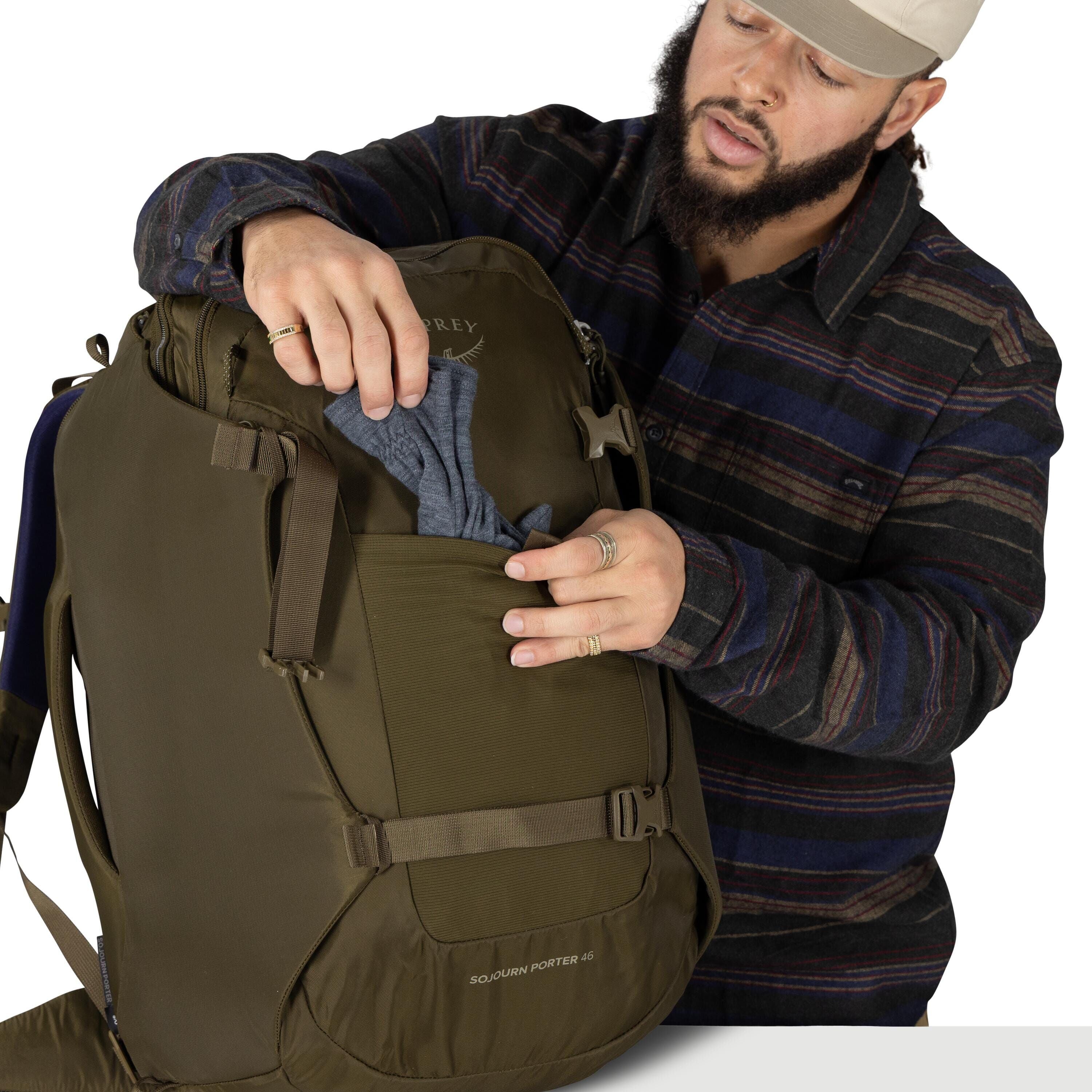 A person wearing a striped flannel shirt and a beige cap is packing an olive-green Osprey backpack. They are placing a gray garment into a front stretch pocket while holding the pocket open with both hands. The pack features compression straps and reads "