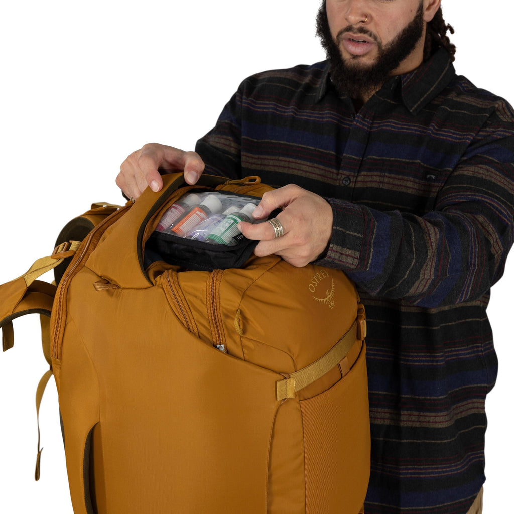 A person opens the top pocket of a mustard-yellow Osprey backpack. Inside the pocket is a clear, zippered pouch containing several small travel bottles with different colored labels. The person wears a dark, striped button-up shirt and silver rings. The s