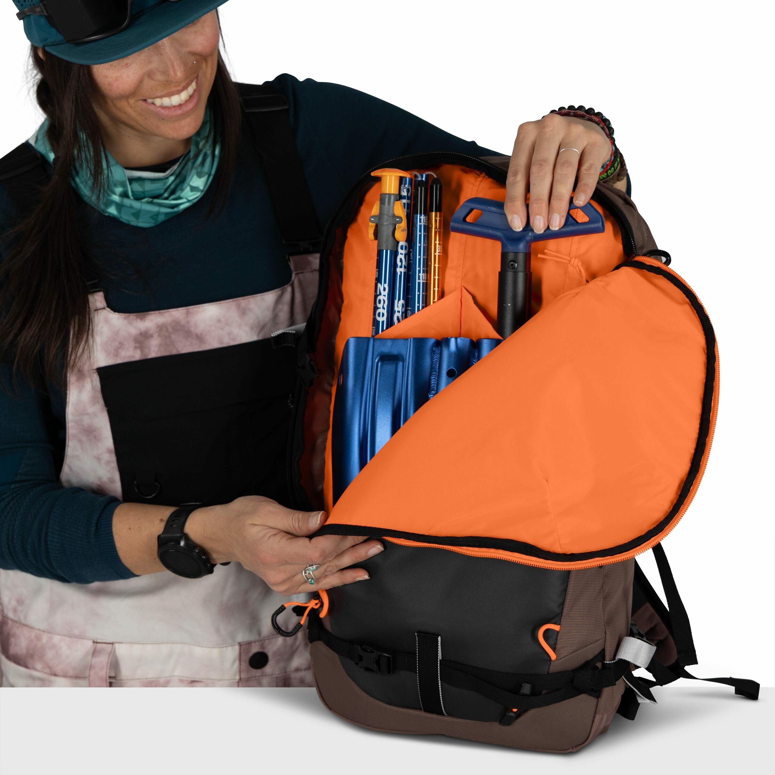 The image shows a person holding open a backpack with an orange interior lining. Inside the backpack, there are several pieces of equipment, including a blue snow shovel and what appear to be avalanche probes, which are tools commonly used for snow and mo