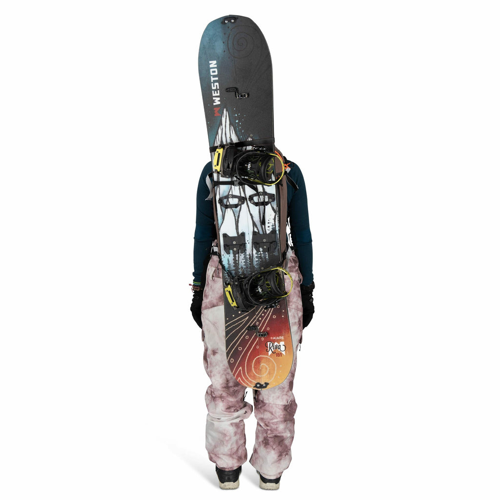 The image features a person dressed in winter sports gear, viewed from the back. They are wearing a teal long-sleeve top, pink and white tie-dye snow pants, and black gloves. A snowboard is strapped vertically to their back, with the brand "Weston" visibl