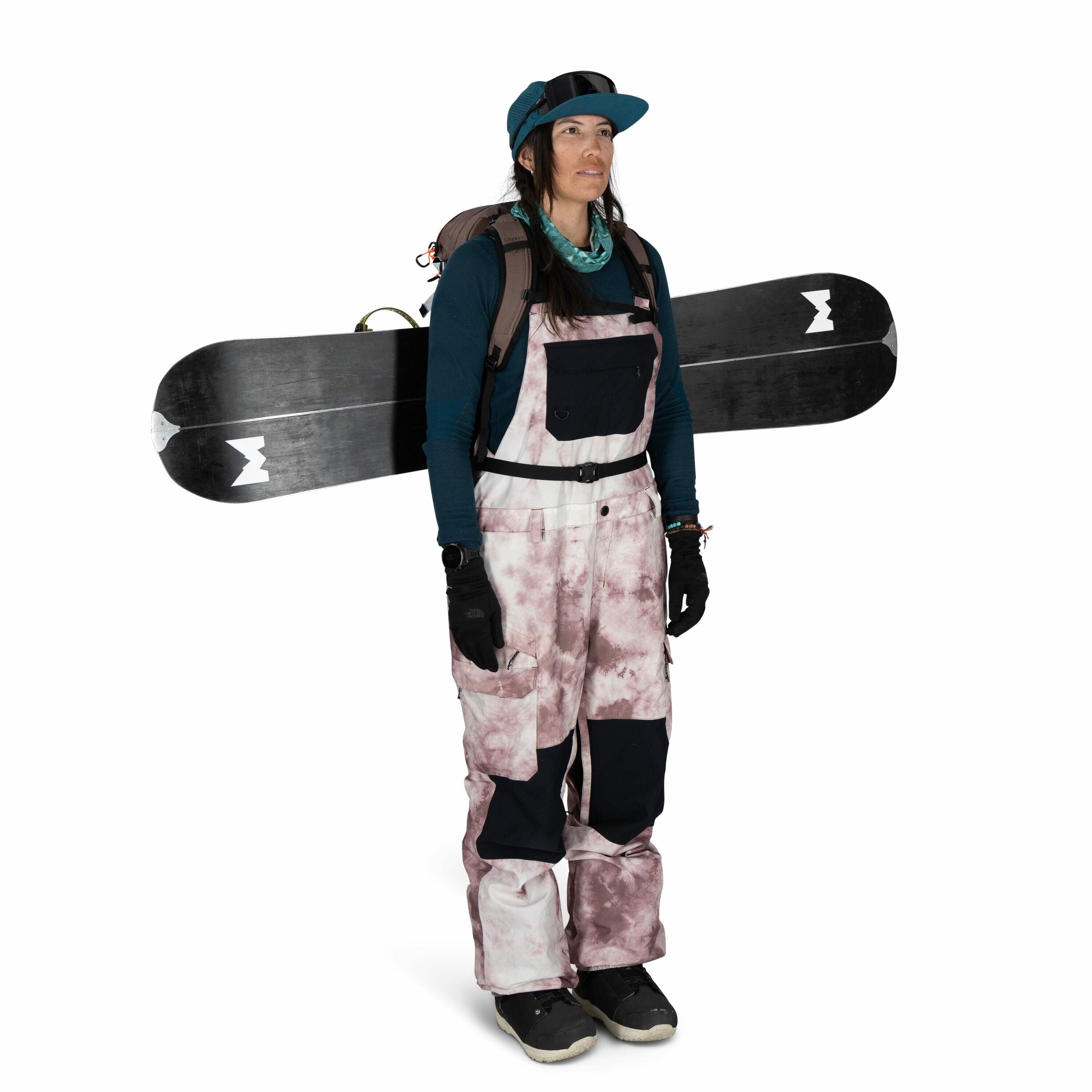 The image features a person prepared for snowboarding. They are wearing a teal beanie, teal gloves, and a long-sleeve dark teal shirt, complementing their outfit which includes snow bib pants with a tie-dye pattern of pink and black. The individual has a