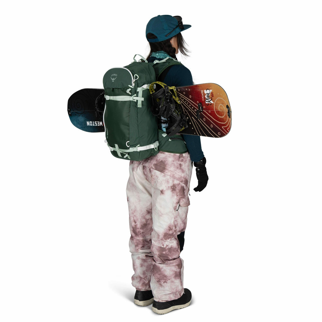 The image features a person dressed in winter gear, standing with their back to the camera. They are wearing a teal baseball cap, a dark-colored jacket, and pinkish-gray patterned snow pants. The person is carrying a large green backpack, to which a snowb