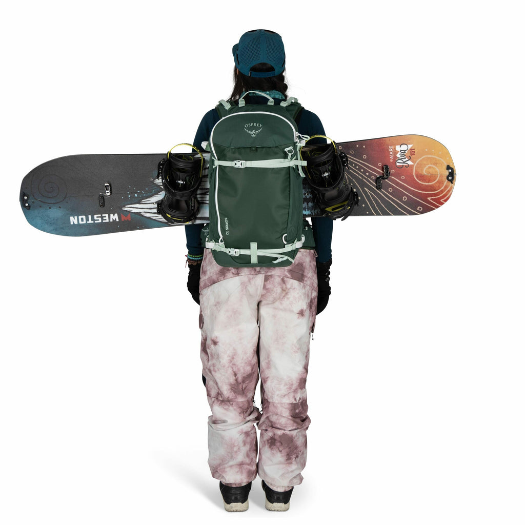 The image shows a person from behind, equipped for snowboarding. They are wearing a teal hat and a teal backpack, which appears to have attachments for carrying a snowboard. The snowboard is mounted horizontally on the backpack and has a vibrant design wi