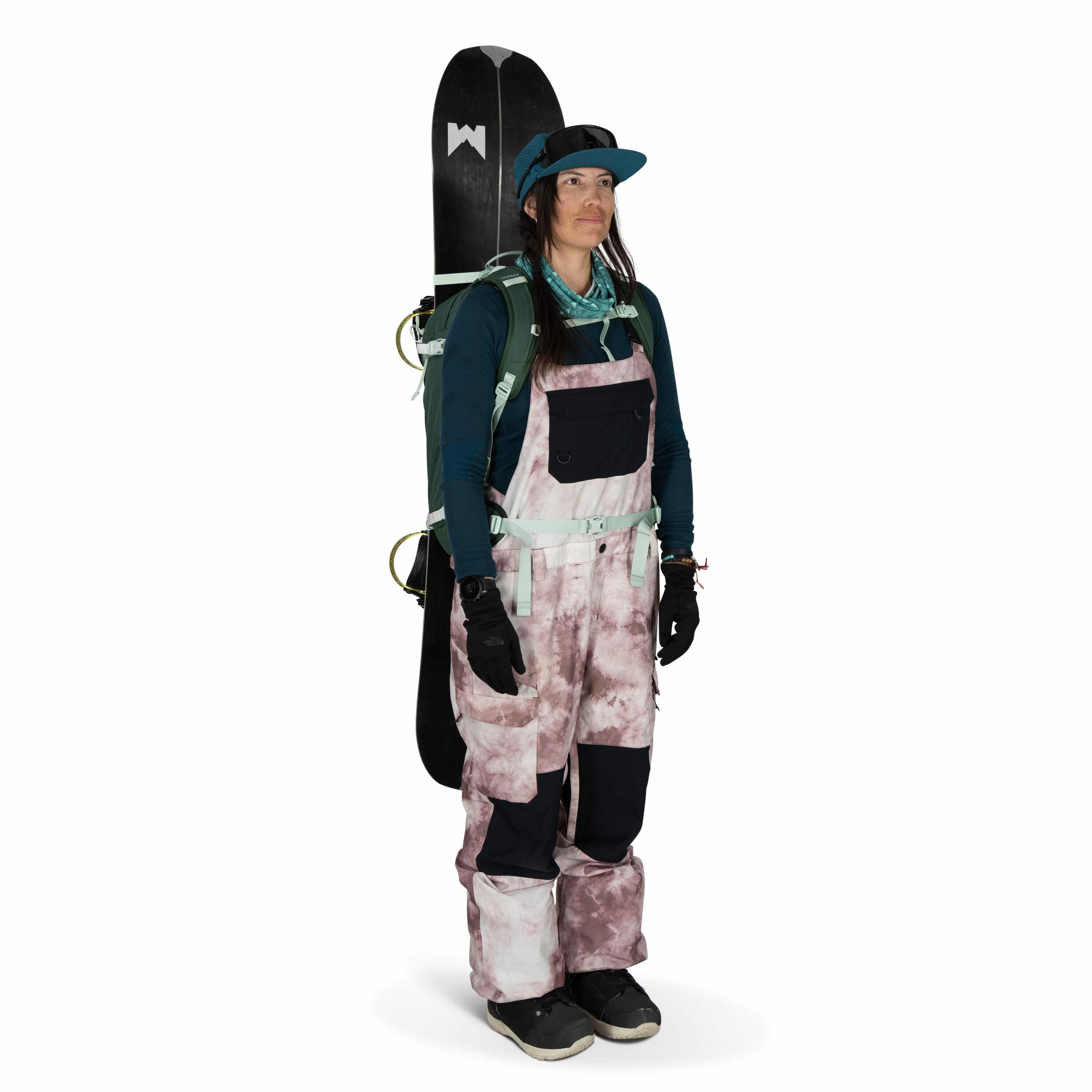 The image shows a person dressed in snowboarding gear. They are wearing a teal long-sleeve shirt under a pair of pink and white patterned bib snow pants with black patches on the knees. The person has on black gloves and black snowboard boots, and they ca