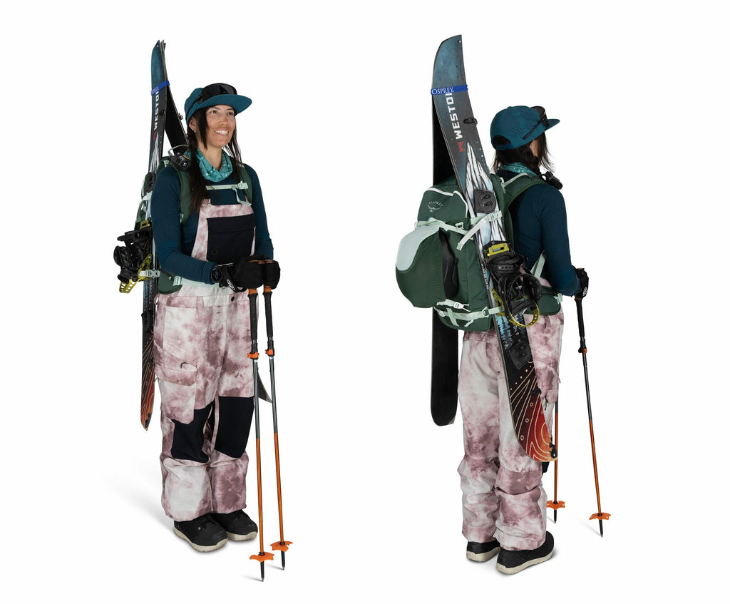 The image shows a person dressed in ski gear, equipped for outdoor adventure in snowy conditions. They are wearing a blue long-sleeve top underneath pink and black patterned overalls. The individual has a large green backpack with attached skis and poles,