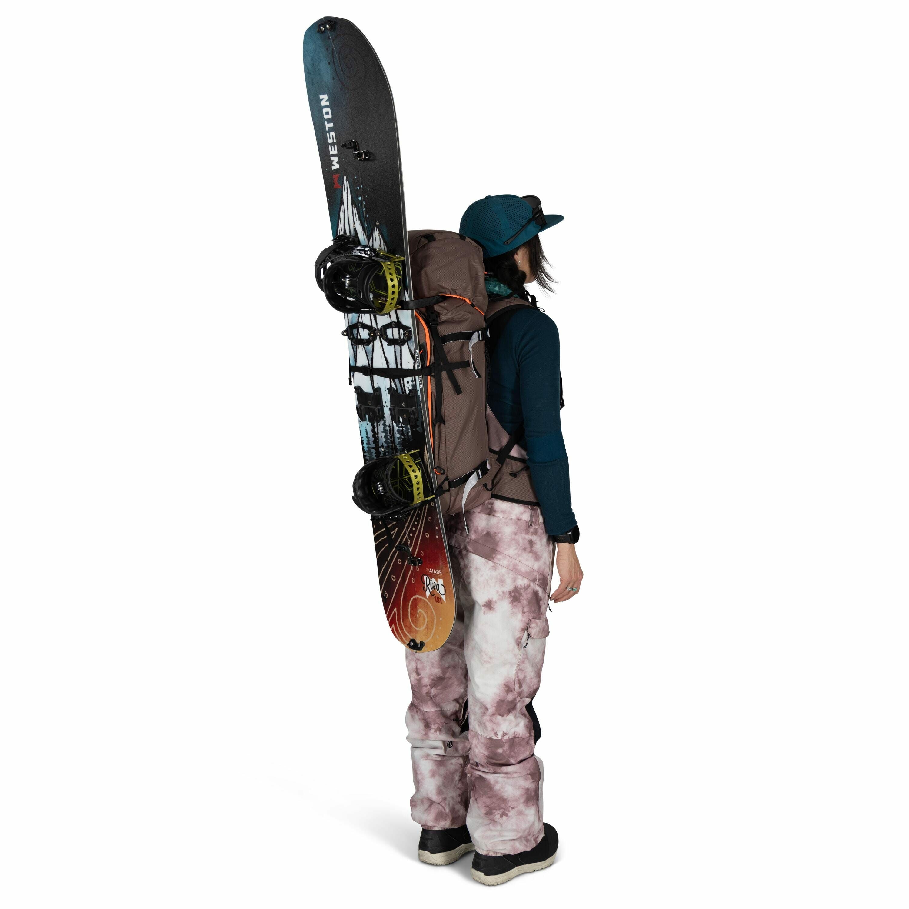 The image features a person standing in a side profile, geared up for snowboarding. They are carrying a snowboard vertically attached to a backpack. The snowboard has a colorful design with a prominent black-and-white mountain illustration and bright red