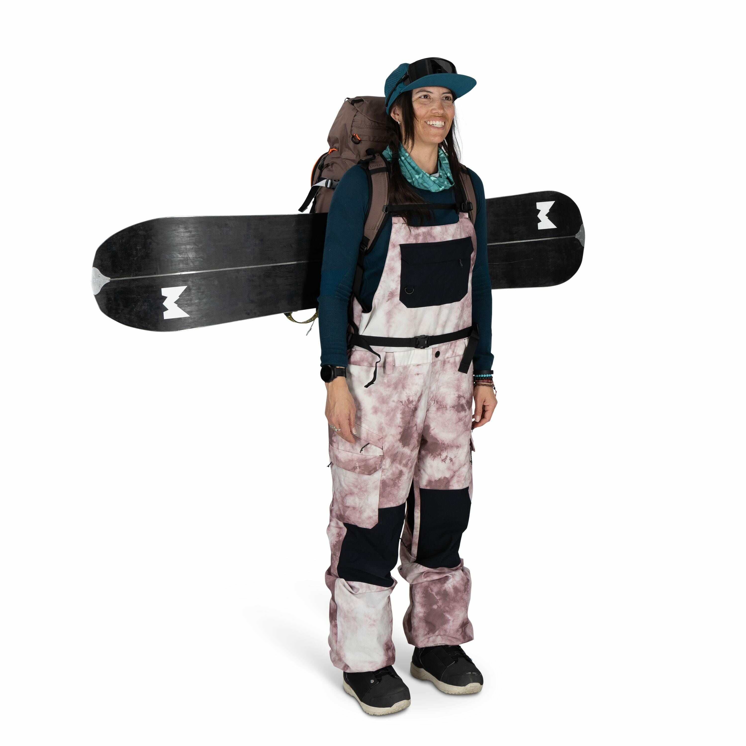 The image shows a person dressed in winter sportswear, likely prepared for snowboarding or skiing. They are wearing a teal long-sleeve top, layered with a pink and white patterned bib-style snow pant, which features black accents on the knees. The individ