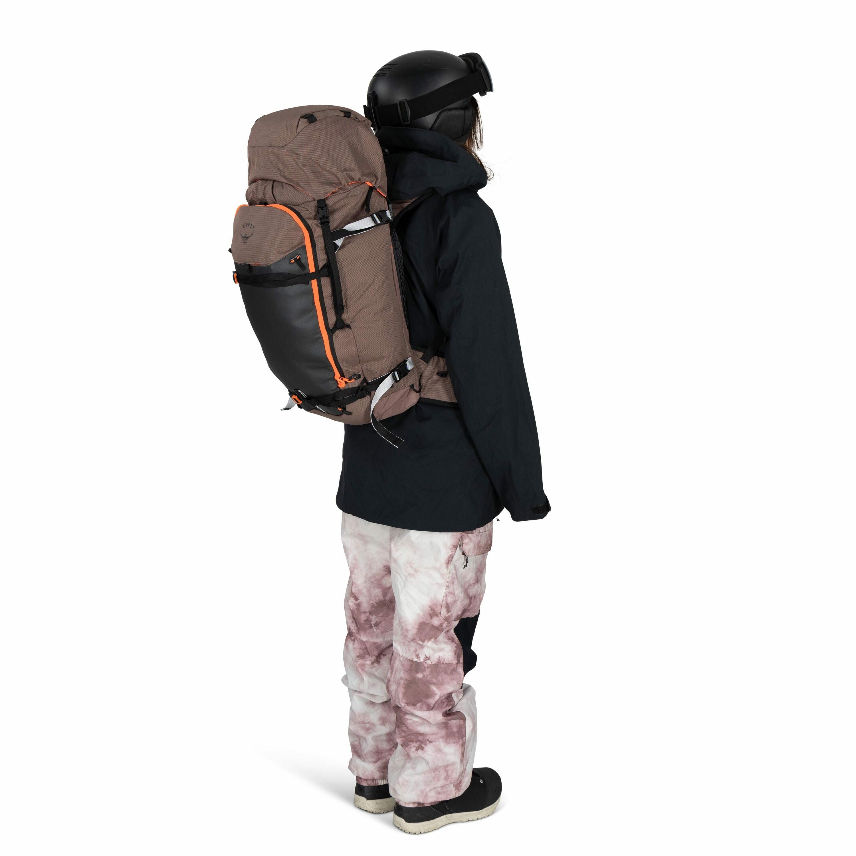 The image shows a person wearing winter outdoor gear, including a large brown and black backpack with orange accents. They are dressed in a black jacket, a black helmet, and goggles, indicating they might be skiing or snowboarding. The individual is also