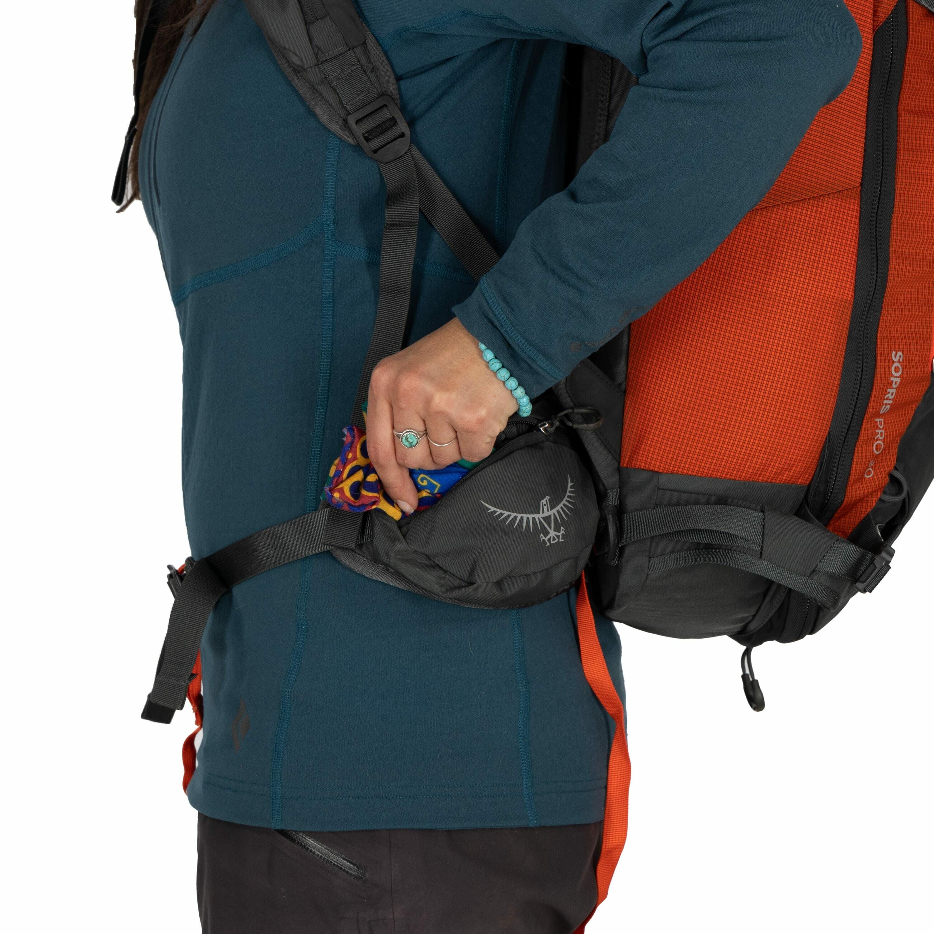 The image shows a person wearing a backpack, with the focus on the side pocket of the backpack. The backpack is a burnt orange color with black accents, and the brand logo "Osprey" is visible on the side pocket. The person is wearing a teal long-sleeve sh