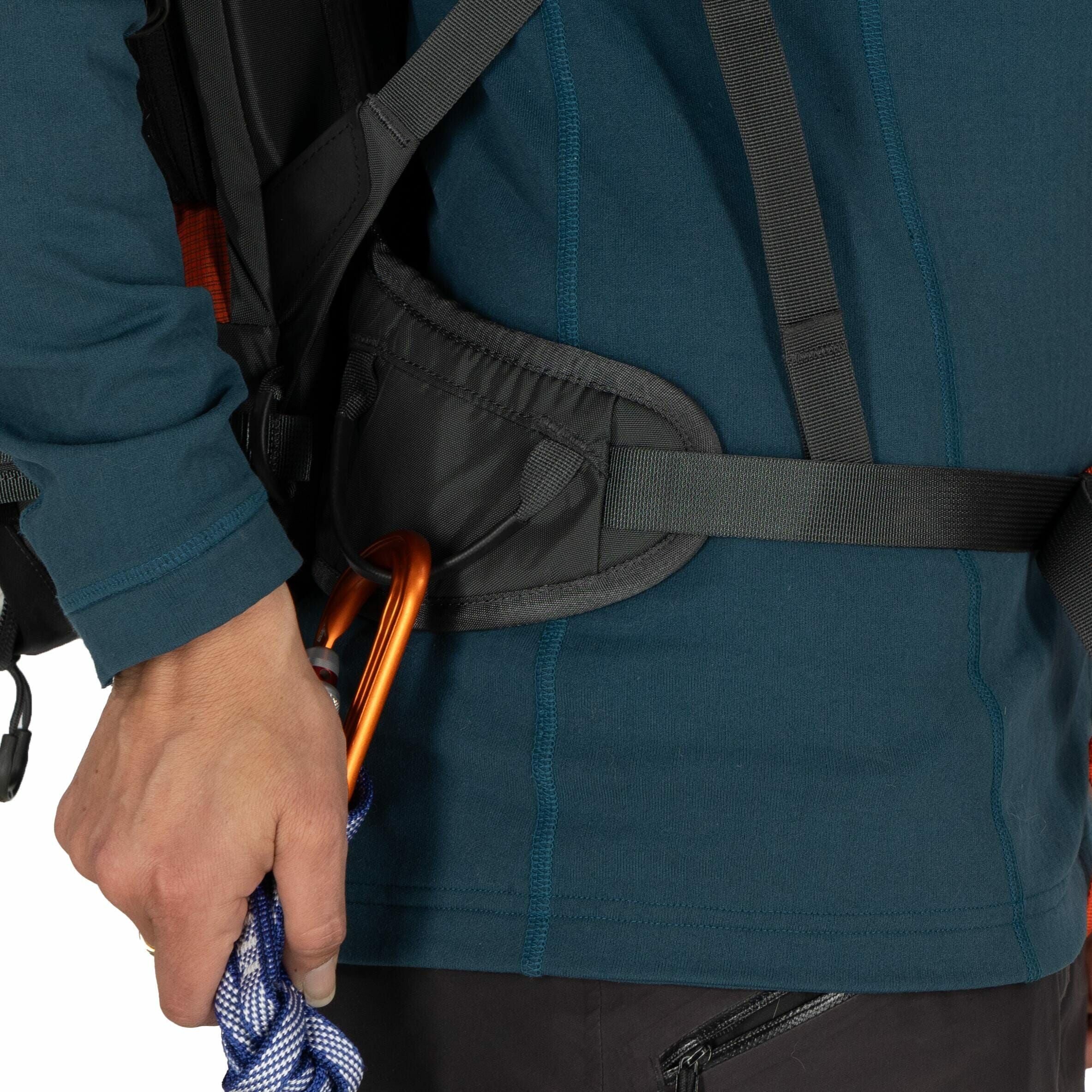 The image shows a person dressed in an outdoor or climbing outfit, with a focus on their lower torso. They are wearing a dark teal long-sleeve top, which has visible seams running vertically and horizontally. The individual has on a backpack with straps a