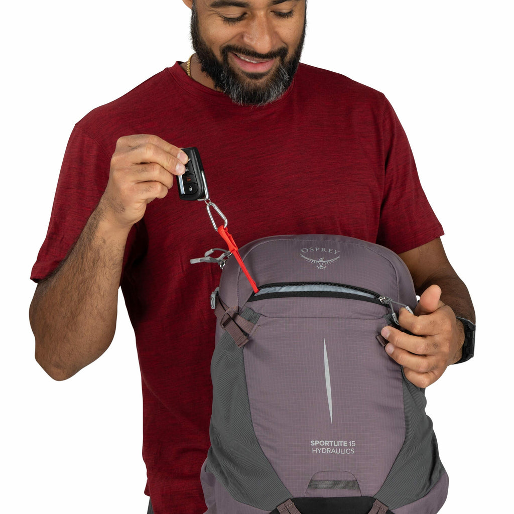 The image shows a person wearing a red t-shirt and holding a gray Osprey Sportlite 15 Hydraulics backpack. The person is using a carabiner with a red strap to attach a set of keys to the backpack. The backpack features several compartments, with one zippe