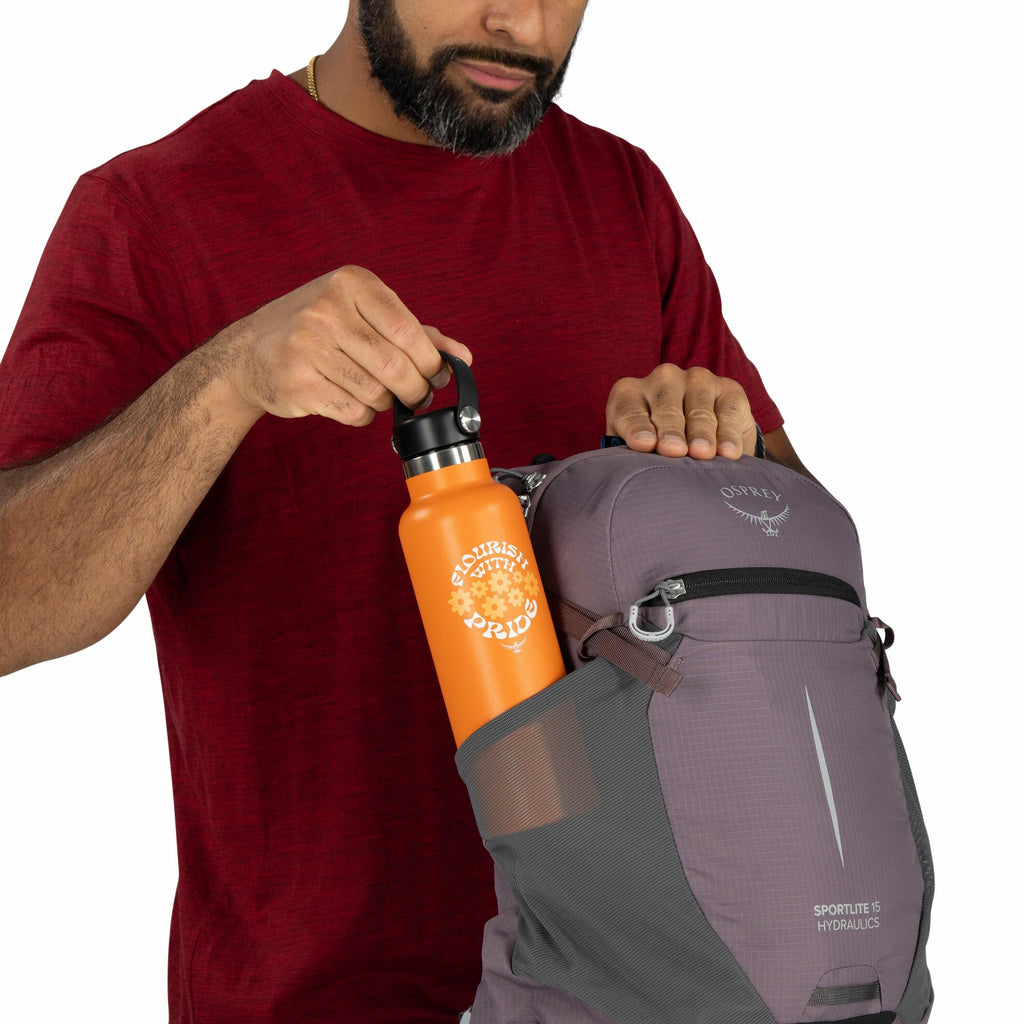 The image shows a man wearing a red shirt and placing an orange water bottle into the side pocket of a grey Osprey backpack, which is labeled "Sportlite 15 Hydraulics." The water bottle features the text "Foolishly With Pride" printed in white, accompanie
