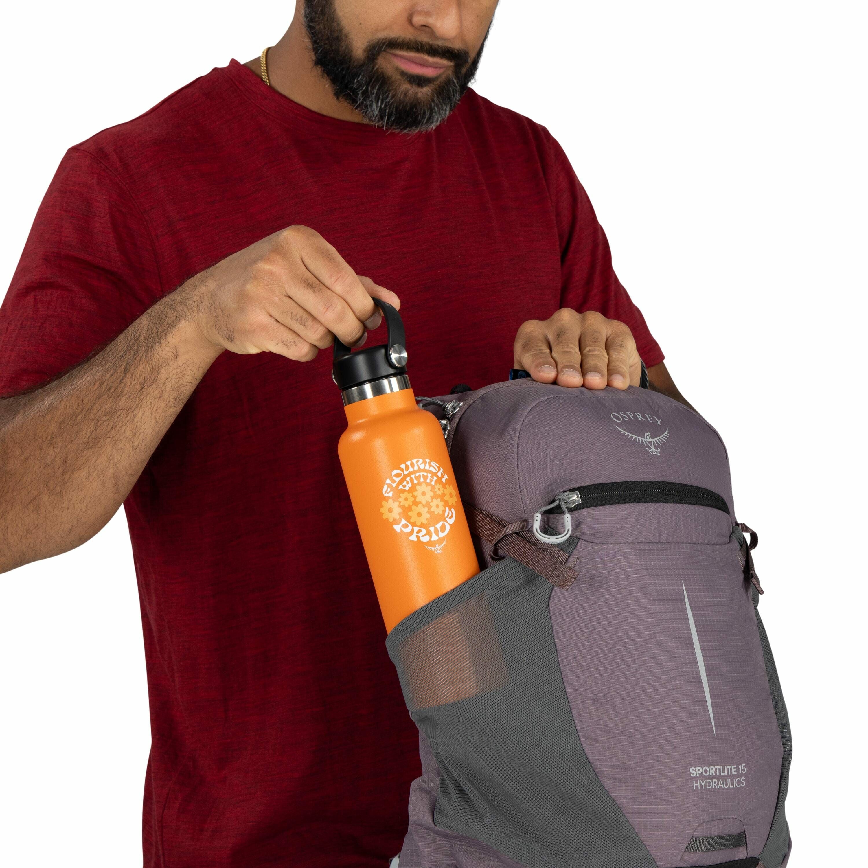 The image shows a man wearing a red shirt and placing an orange water bottle into the side pocket of a grey Osprey backpack, which is labeled "Sportlite 15 Hydraulics." The water bottle features the text "Foolishly With Pride" printed in white, accompanie