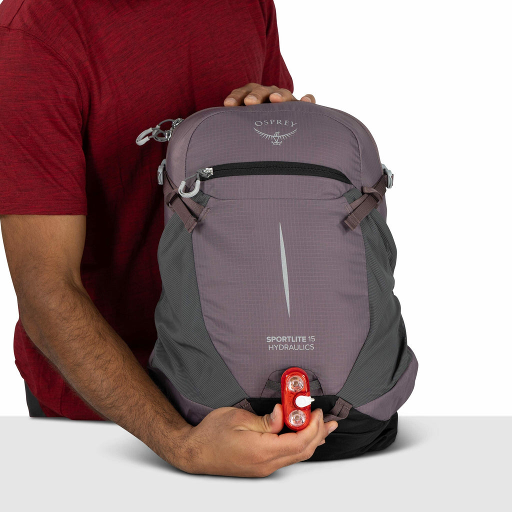 The image shows a person wearing a red shirt holding a backpack with both hands. The backpack is primarily gray with a purple front panel and features the "Osprey" logo at the top, along with the words "Sportlite 15 Hydraulics" printed on the front. A sma