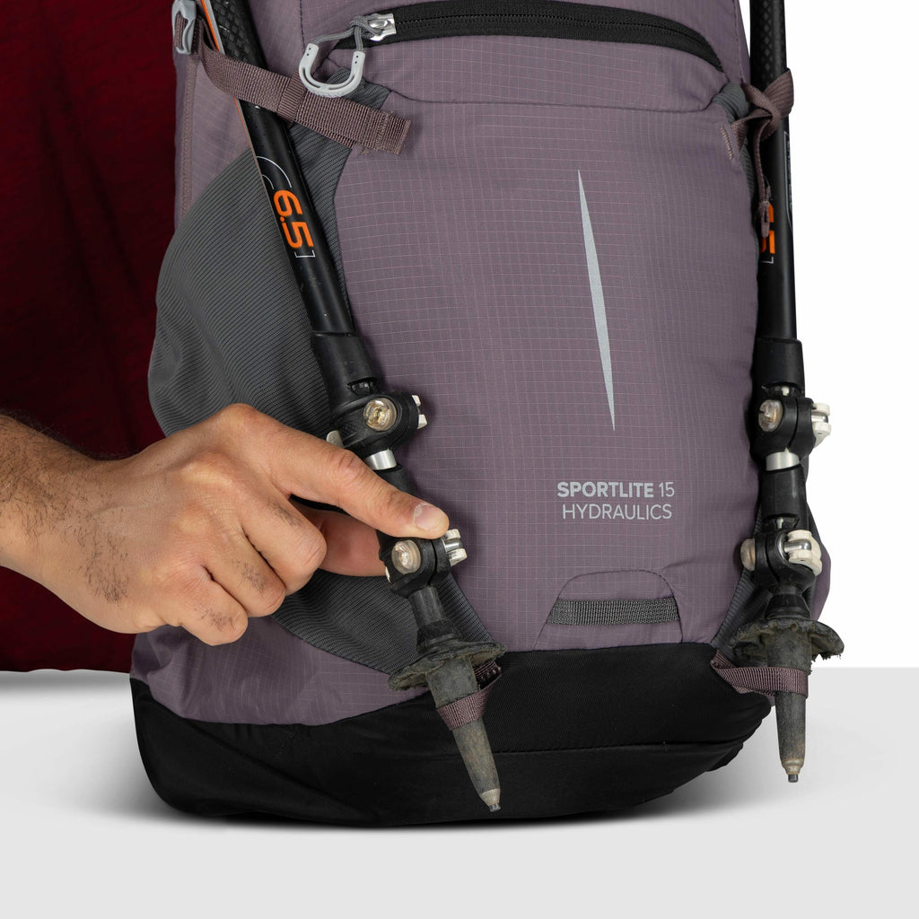 The image shows a close-up of a backpack with a purple and black color scheme. The backpack has the logo "SPORTLITE 15 HYDRAULICS" printed on its front. Two trekking poles are attached to the front of the backpack using small black clips, and a person's h