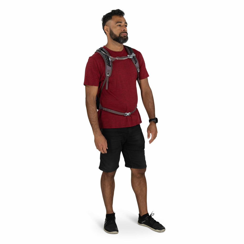 The image shows a person standing casually, wearing a red t-shirt and black shorts. They have a gray backpack with multiple straps over their shoulders. The individual is also wearing black athletic shoes and a watch on their left wrist. The background is