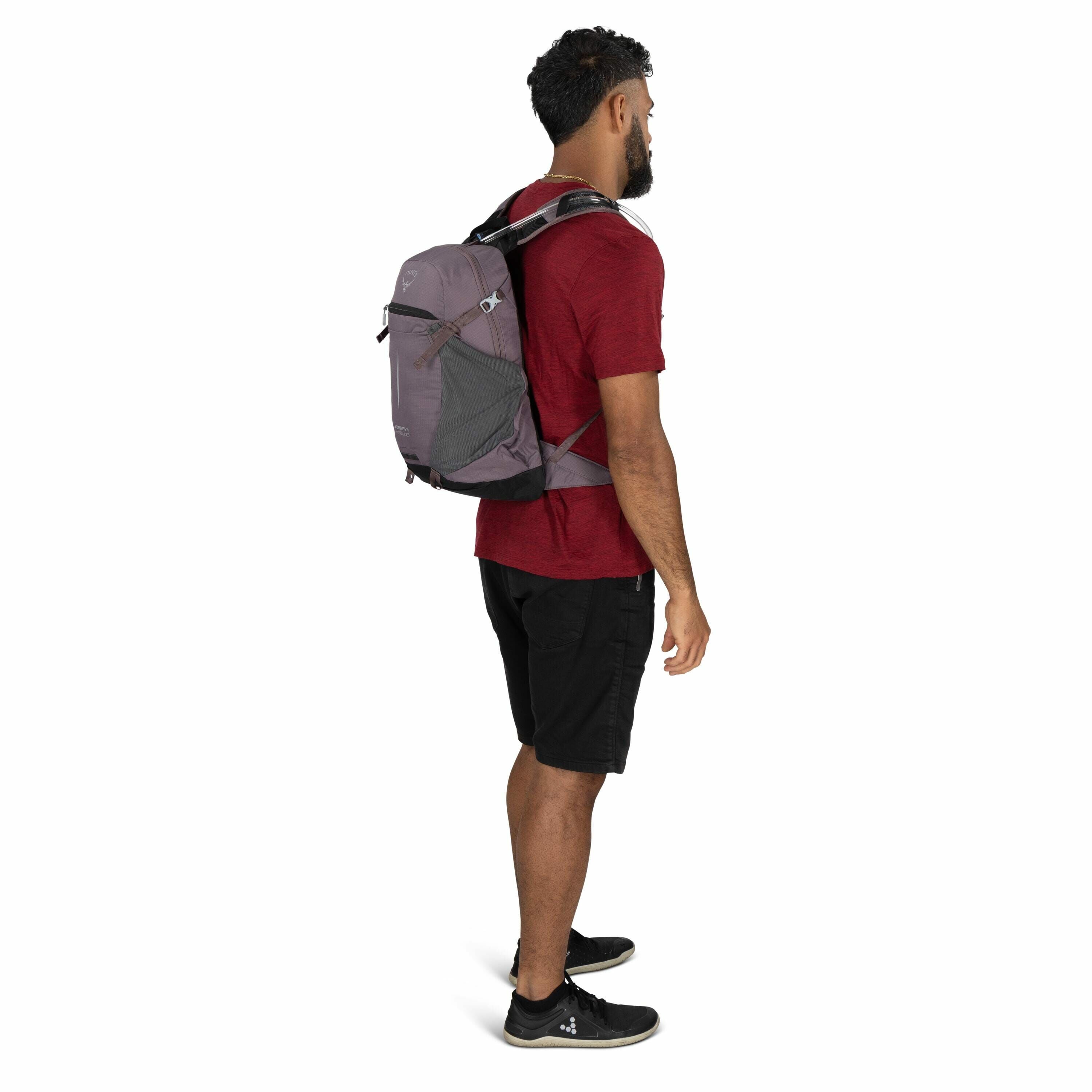The image shows a person standing in a side profile, wearing a red short-sleeve t-shirt and black shorts. They have a backpack from the brand Osprey strapped securely on their back. The backpack appears to be designed for outdoor or hiking use, given its