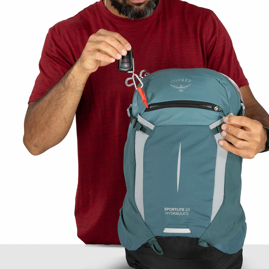 The image shows a man wearing a red shirt holding a blue Osprey backpack, which is part of the Sportlite 20 Hydraulics series. The man is placing a set of car keys, attached to a red clip, into a small zippered pocket on the front of the backpack. The Osp