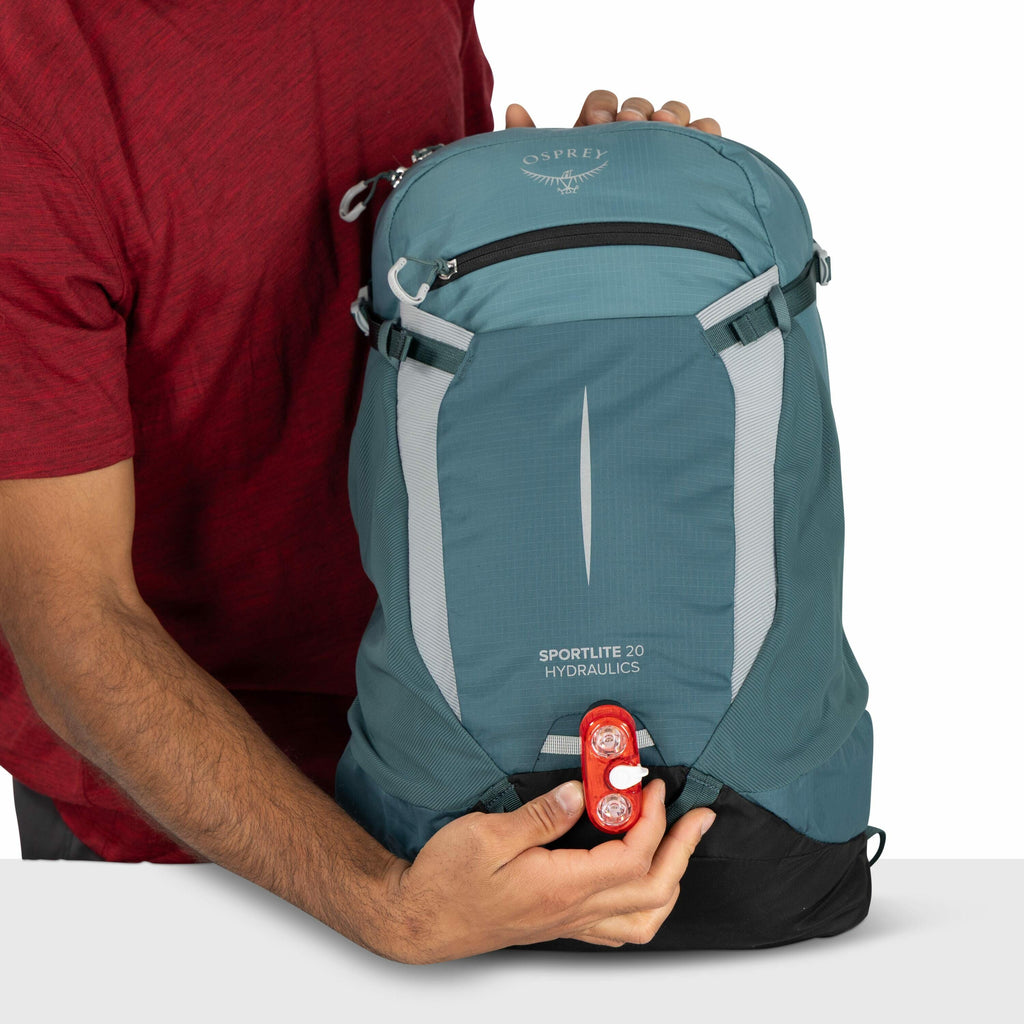 The image shows a person in a red shirt holding a blue Osprey backpack. The model of the backpack is indicated as "Sportlite 20 Hydraulics," which is written on the front. The person is attaching or detaching a red safety light to the lower part of the ba