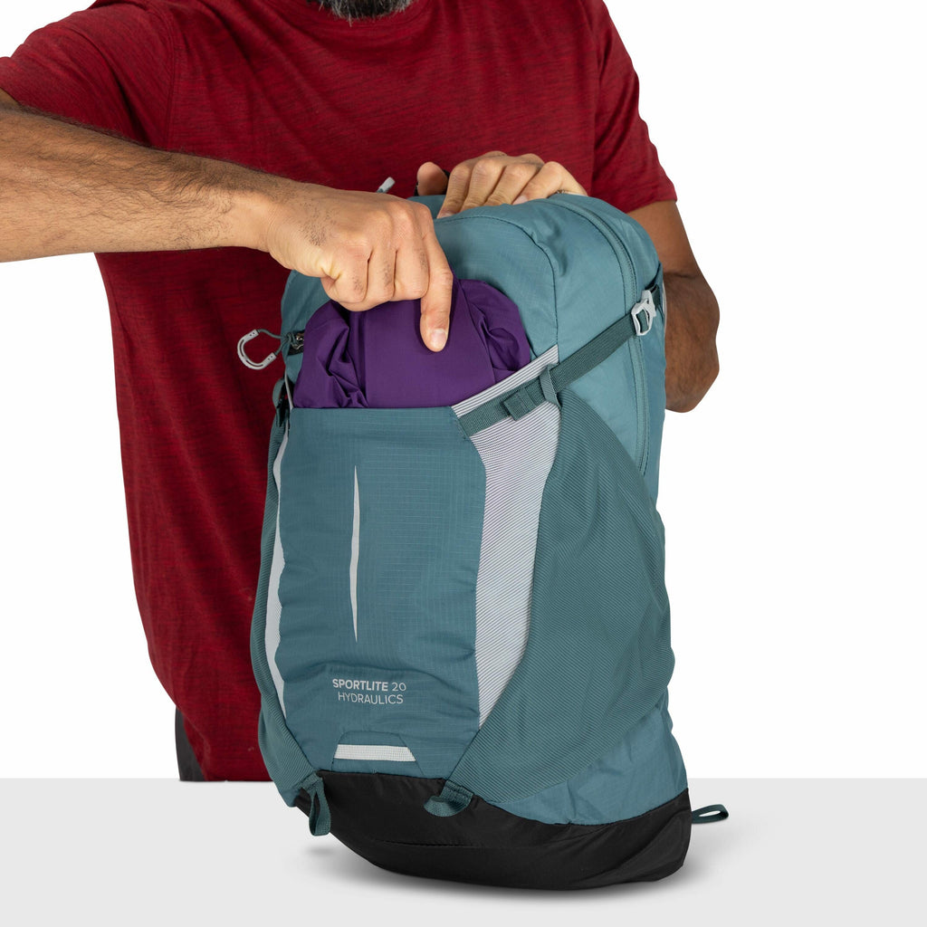 The image depicts a person wearing a red shirt placing a purple item into the front pocket of a teal and black backpack. The backpack has the text "SPORTLITE 20 HYDRAULICS" printed on its front. The person's left arm is visible, showing some beard and arm
