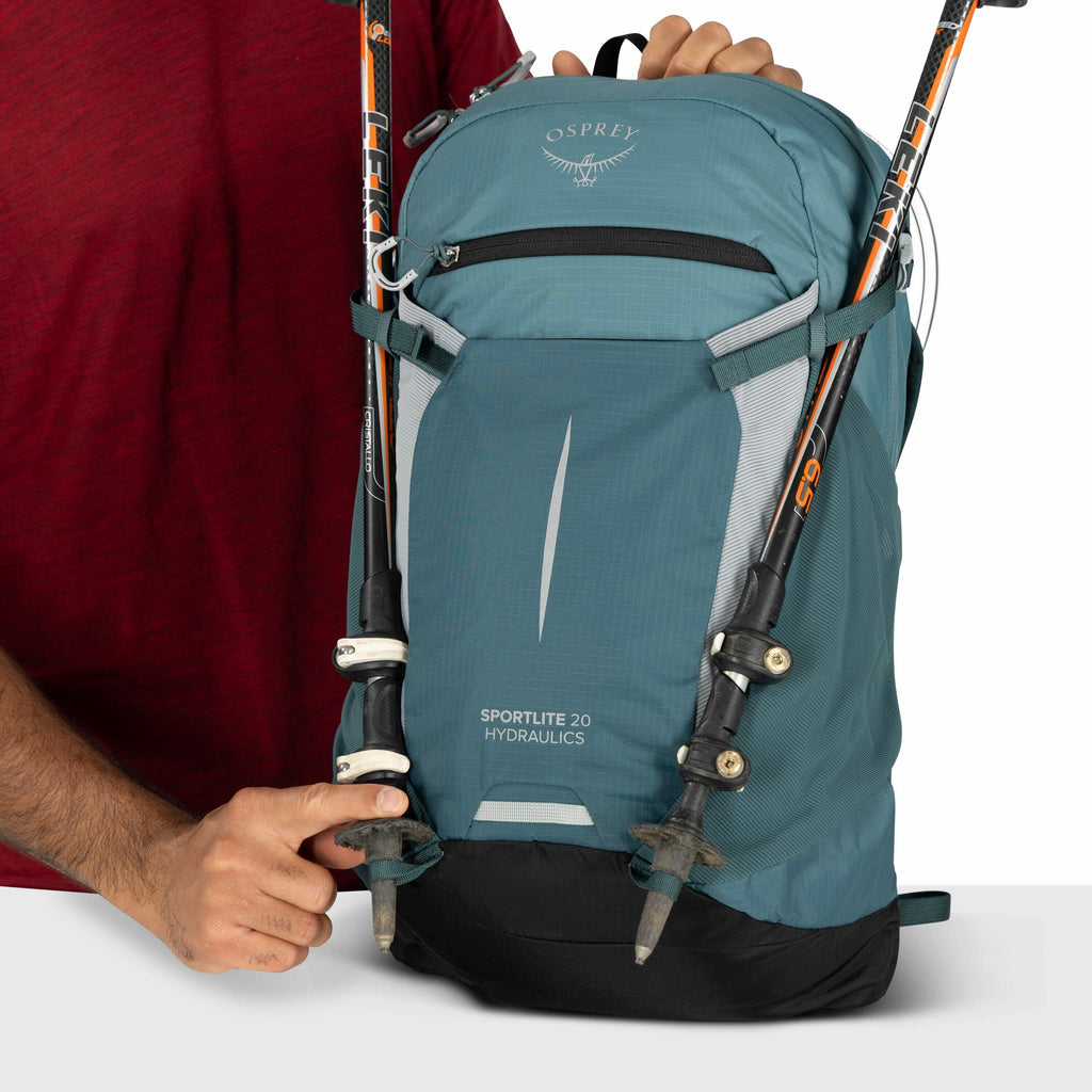 The image shows a person wearing a red shirt holding a blue and black Osprey backpack. The backpack has the model name "SPORTLITE 20 HYDRAULICS" printed on it. Two trekking poles are attached to the front of the backpack, with the brand "LEKI" visible on