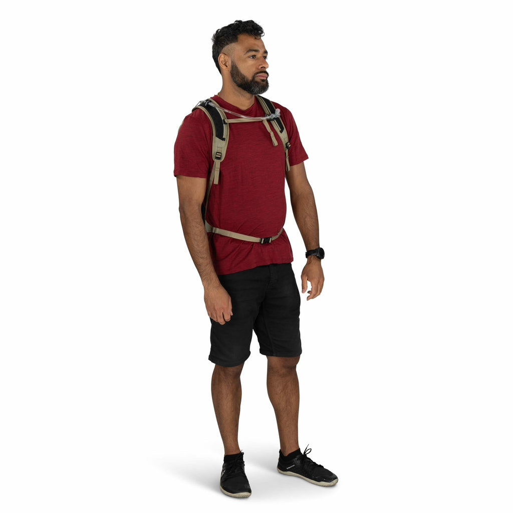 The image shows a man standing against a plain white background. He is wearing a casual outfit consisting of a red short-sleeve T-shirt, black shorts, and black athletic shoes. He has a gray and black backpack, and a black digital wristwatch on his left w