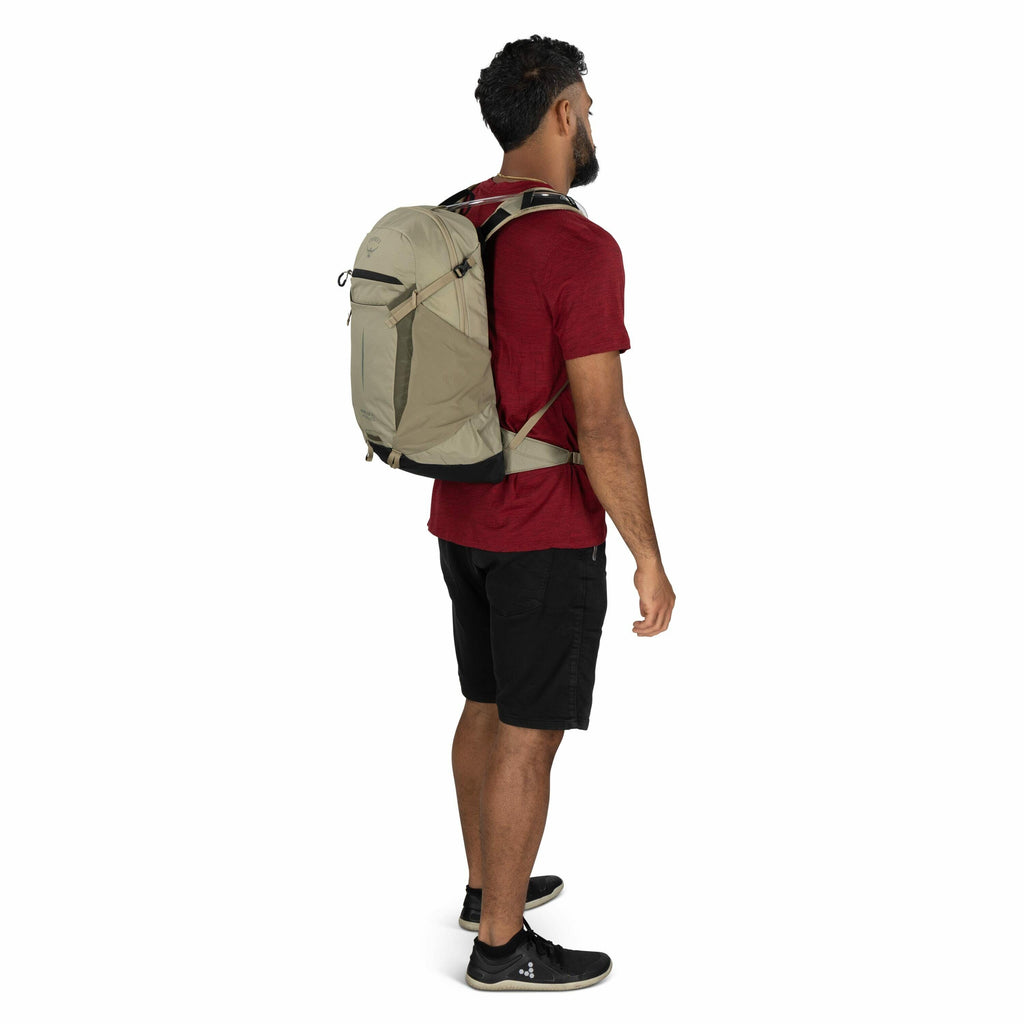 The image depicts a man standing sideways, showcasing a light olive backpack he is wearing. He is dressed in a red t-shirt, black shorts, and black sneakers. The backpack features a logo on the top flap, a front zipper pocket, and adjustable straps with a