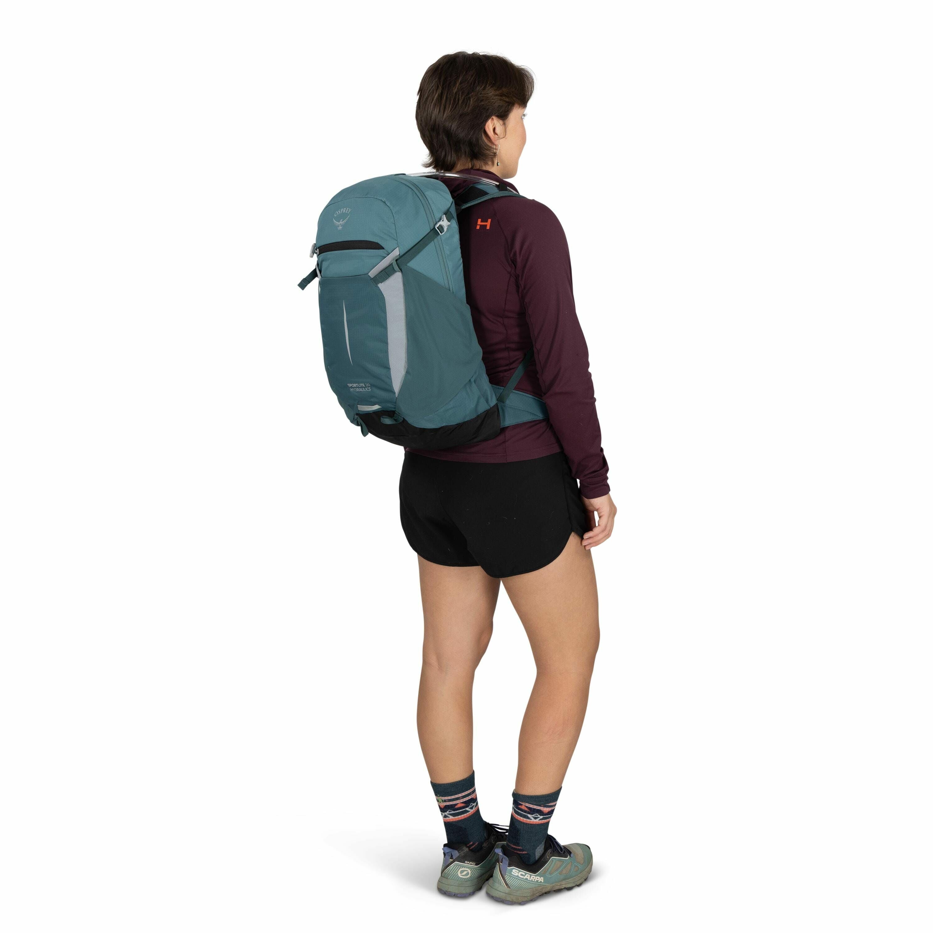 The image depicts a person standing with a large teal-colored Osprey backpack on their back. The individual is wearing a maroon long-sleeve top with a visible small orange "H" logo on the upper left side of the back. They are also dressed in black shorts
