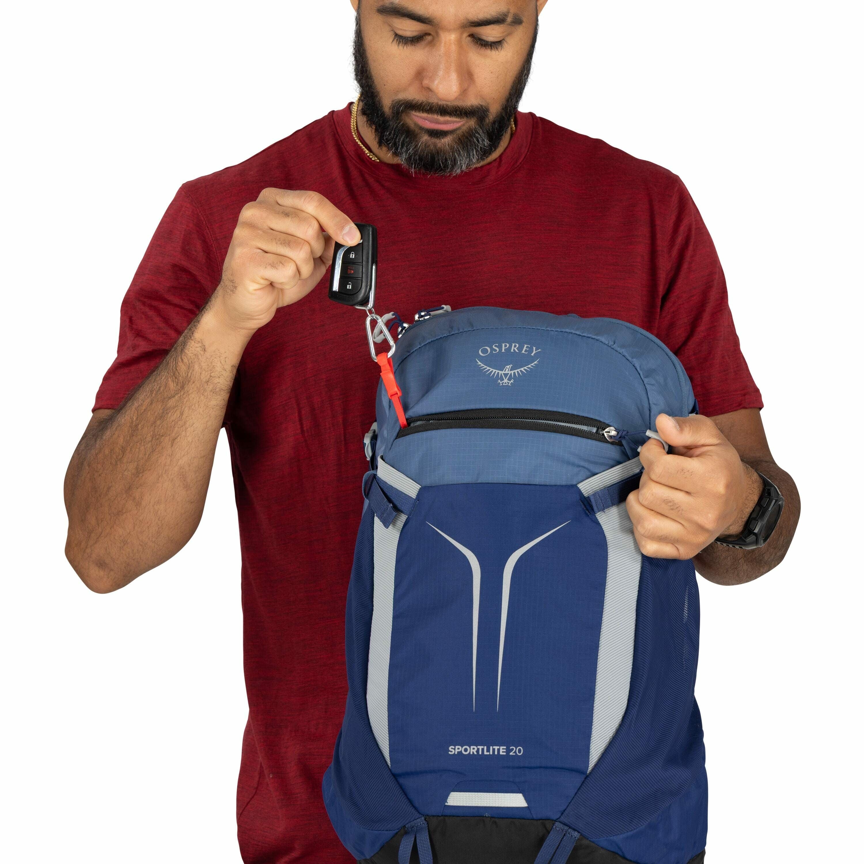 The image shows a person wearing a red t-shirt and holding a blue and gray Osprey backpack, specifically the Sportlite 20 model. The individual is also holding a set of car keys attached to a red carabiner clip, which is fastened to the backpack. The Ospr