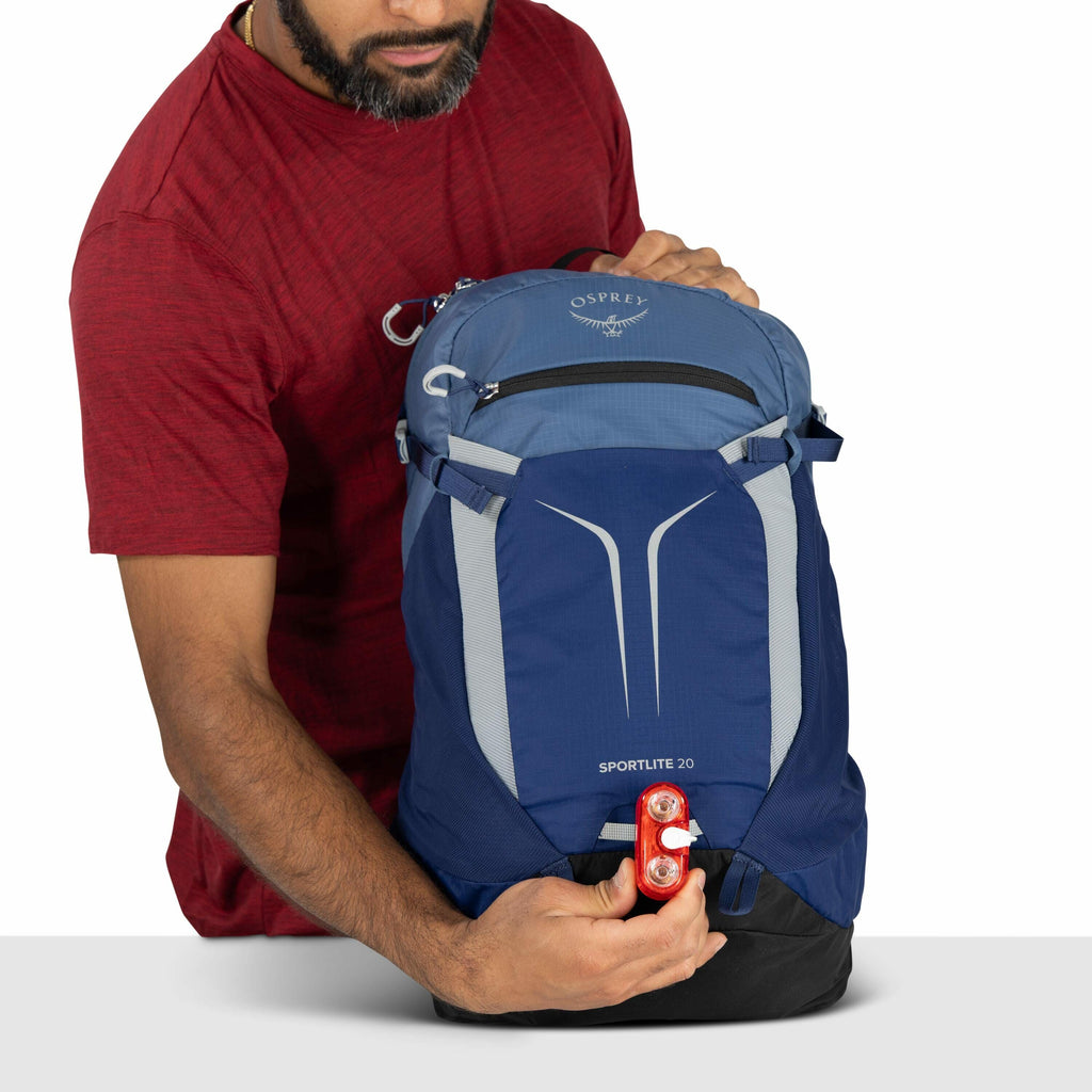 The image shows a man wearing a red shirt holding a blue and light blue backpack. The backpack is branded with the "Osprey" logo on the top pocket and is labeled "Sportlite 20" on the front. The man is attaching or holding a small red light at the bottom