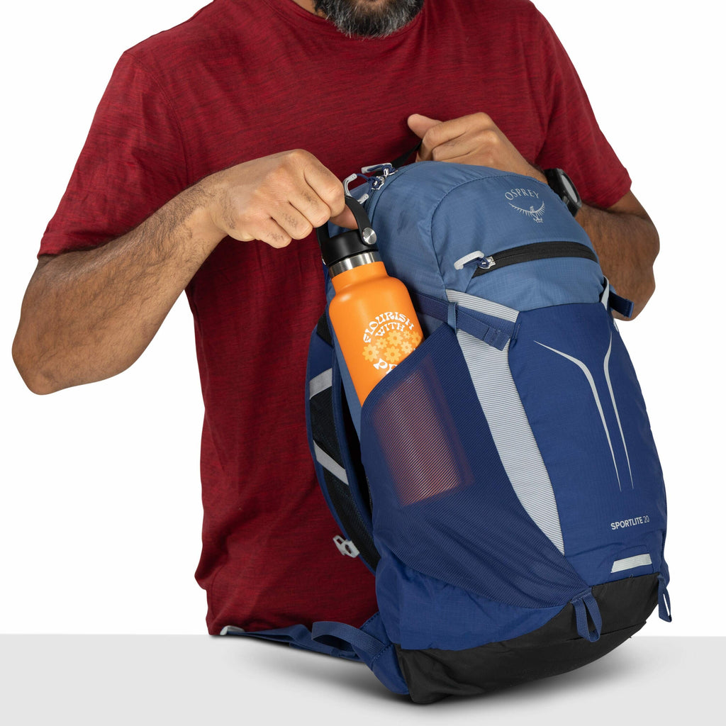The image depicts a man wearing a red shirt and a wristwatch, holding a backpack with both hands. He is inserting an orange water bottle with a black lid into the side mesh pocket of the backpack. The backpack is predominantly blue with light gray and bla