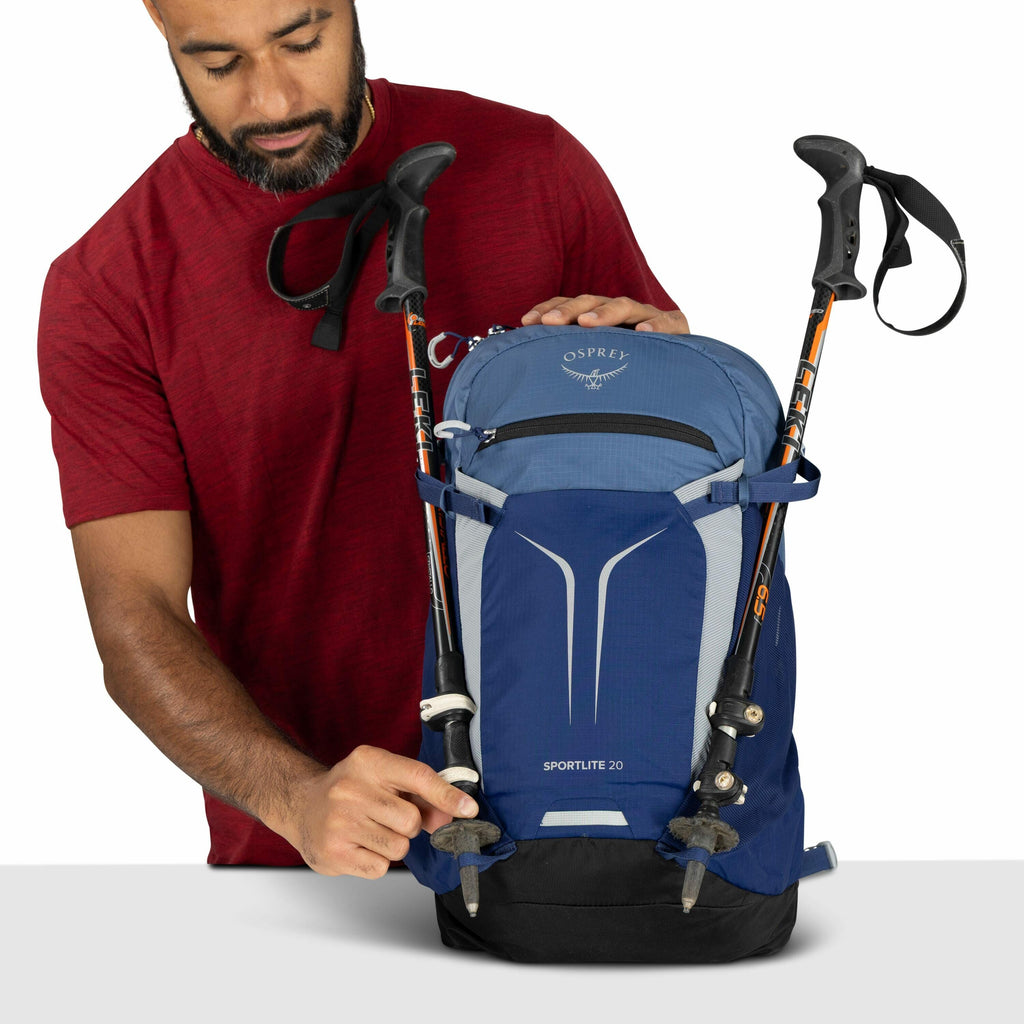 The image features a person holding a blue hiking backpack made by Osprey. The backpack, labeled "SPORTLITE 20," is equipped with two trekking poles that are secured on the front using dedicated straps. The person is wearing a red short-sleeve shirt and i