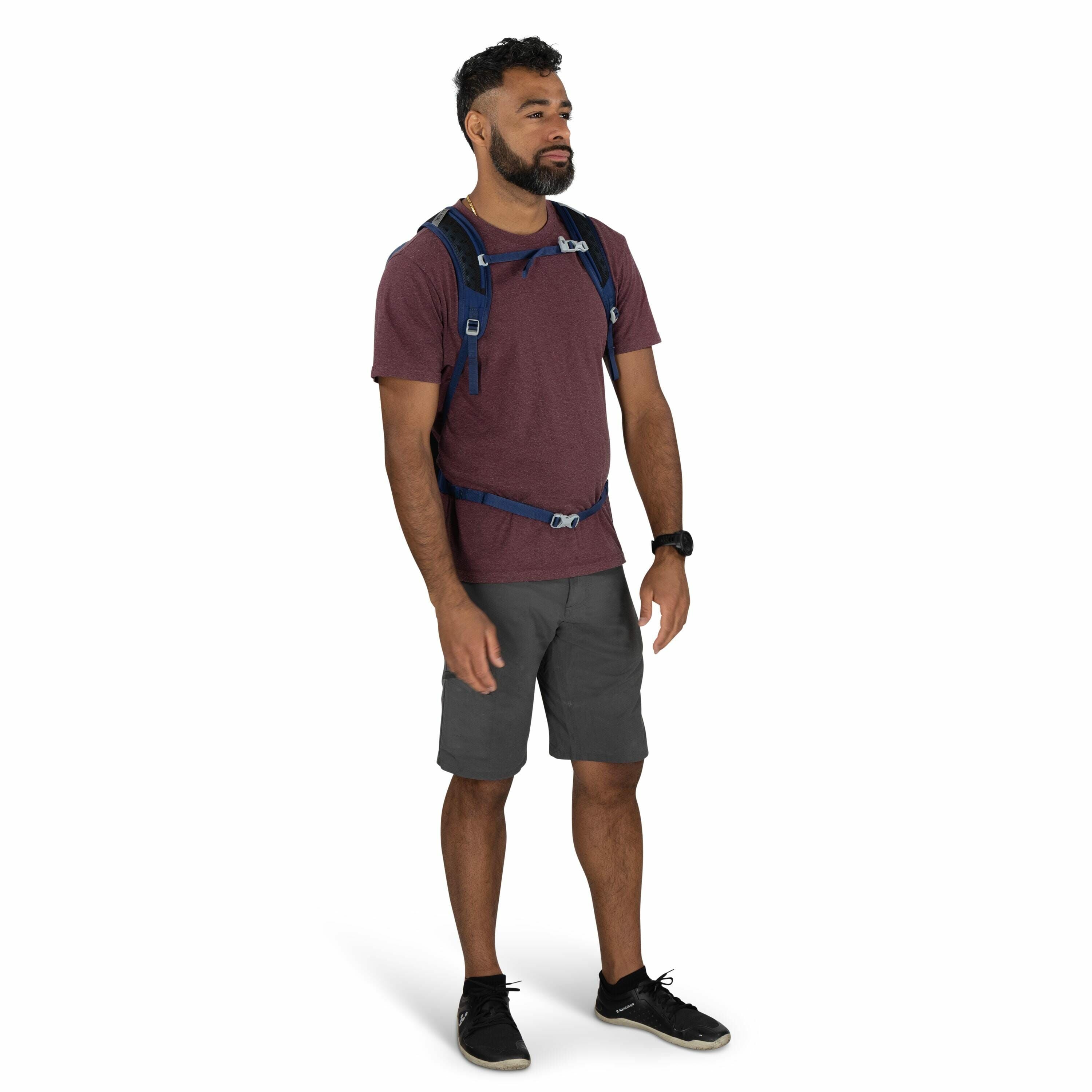 The image shows a man standing in a relaxed pose, wearing outdoor casual clothing suitable for a hike or a casual day out. He is wearing a maroon short-sleeve T-shirt paired with dark gray shorts, along with black sneakers featuring white soles. The man h