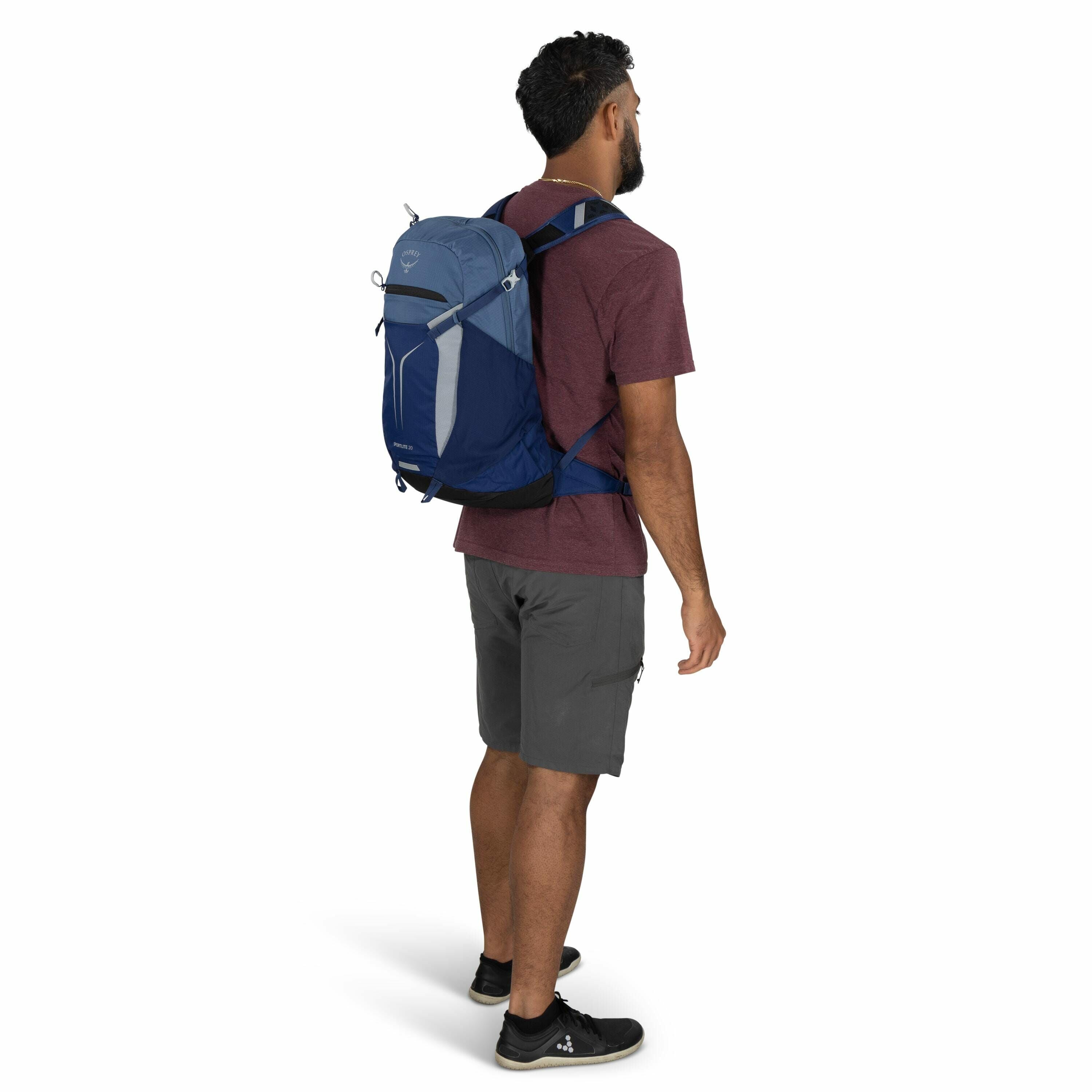 The image features a man standing and facing slightly to the side, wearing a casual outdoor outfit consisting of a maroon t-shirt, gray cargo shorts, and black sneakers. He is carrying a blue and light blue Osprey backpack on his back, which appears to be