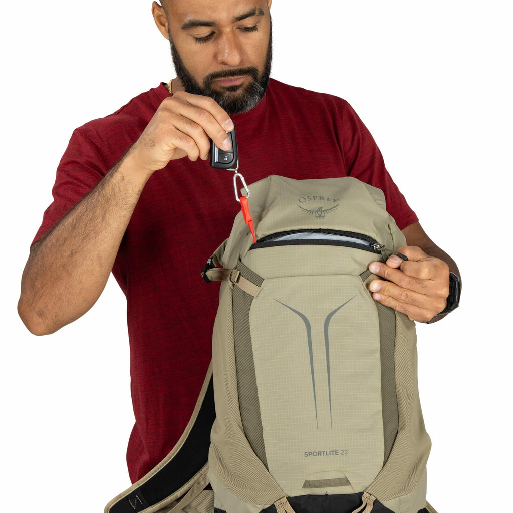 The image shows a person holding a tan-colored backpack in front of a white background. The backpack is branded with the Osprey logo and the model name "Sportlite 22." The individual is wearing a red t-shirt and a black wristwatch and is holding a set of