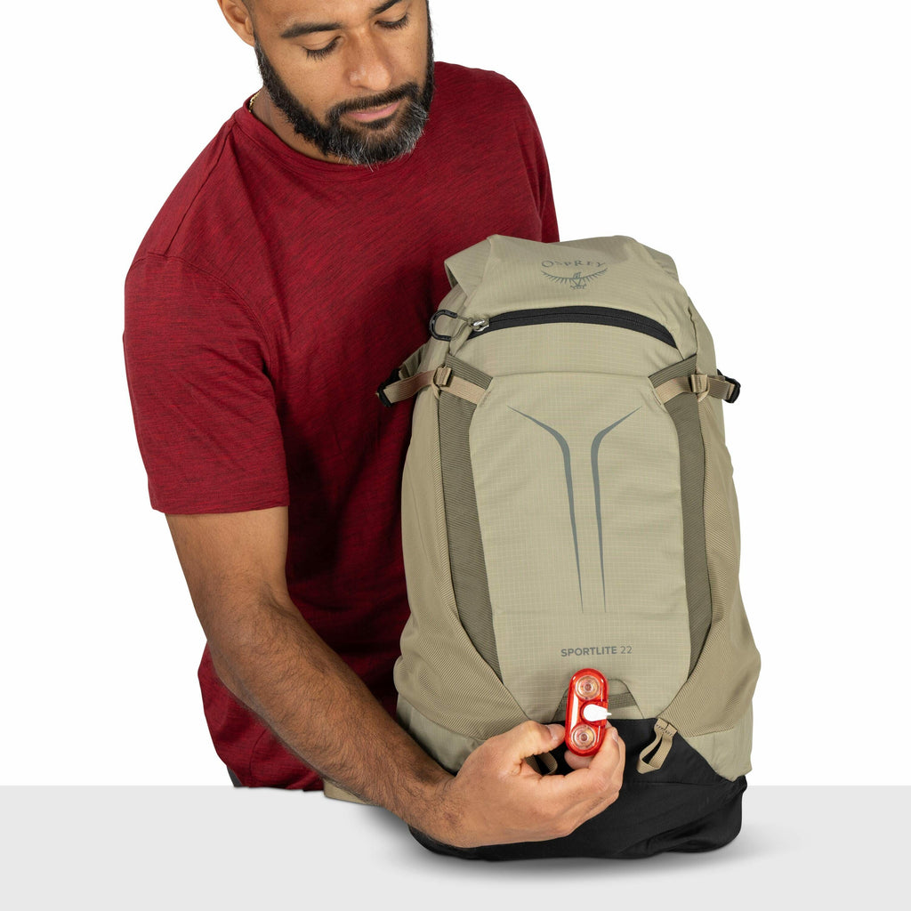 The image shows a person wearing a red shirt and holding a backpack. The backpack is primarily gray with black accents and features the Osprey logo at the top. The model is called "Sportlite 22," as indicated on the front of the backpack. The person is at