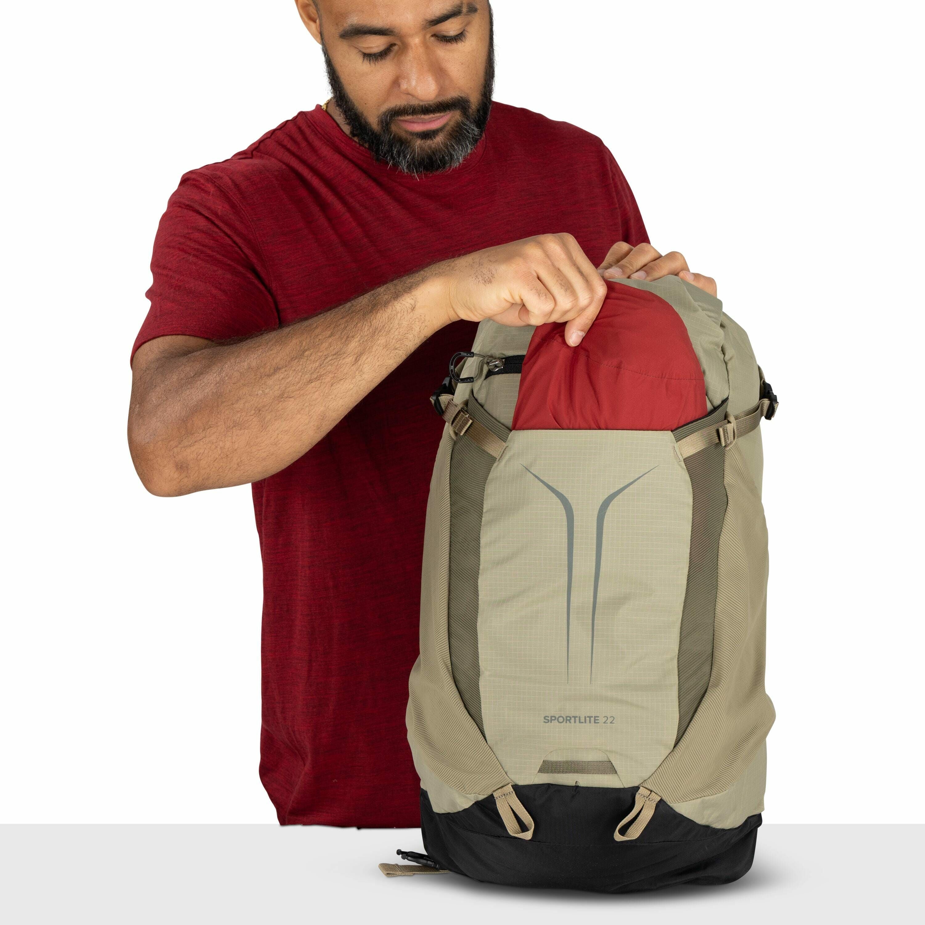 The image shows a person wearing a red short-sleeved shirt, holding a backpack. The backpack is a two-tone design, mainly in beige and light green with accents of black at the bottom. It features the logo "SPORTLITE 22" on the front, indicating the model