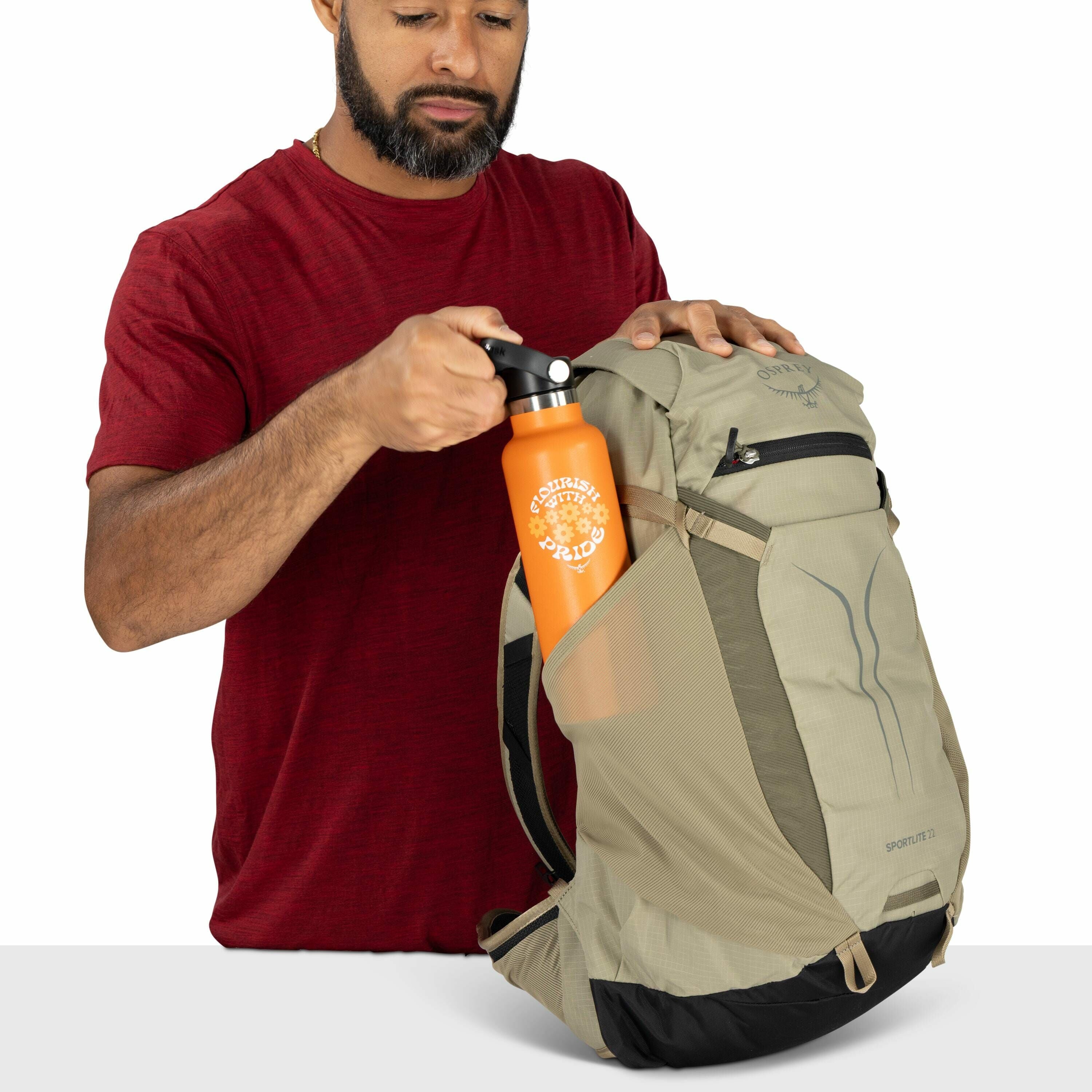 The image depicts a person wearing a red shirt while holding a light tan and gray backpack branded with the "Osprey" logo. The backpack features a front zippered pocket and a side mesh pocket designed for holding items like water bottles. The individual i