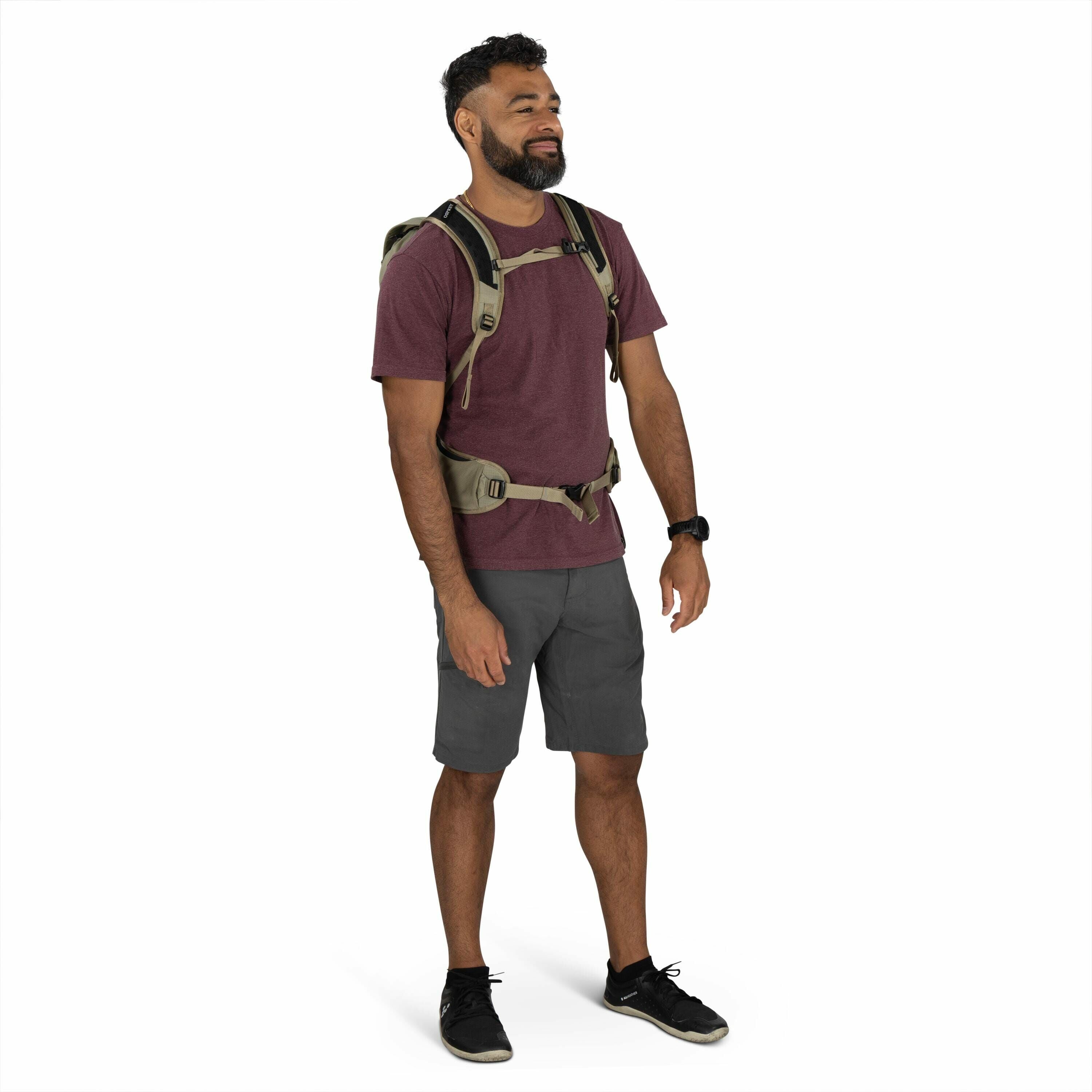 The image shows a man standing against a plain white background. He is wearing a burgundy t-shirt paired with dark gray shorts and black shoes. The man is equipped with an olive-green tactical-style backpack that has multiple straps, including a chest and