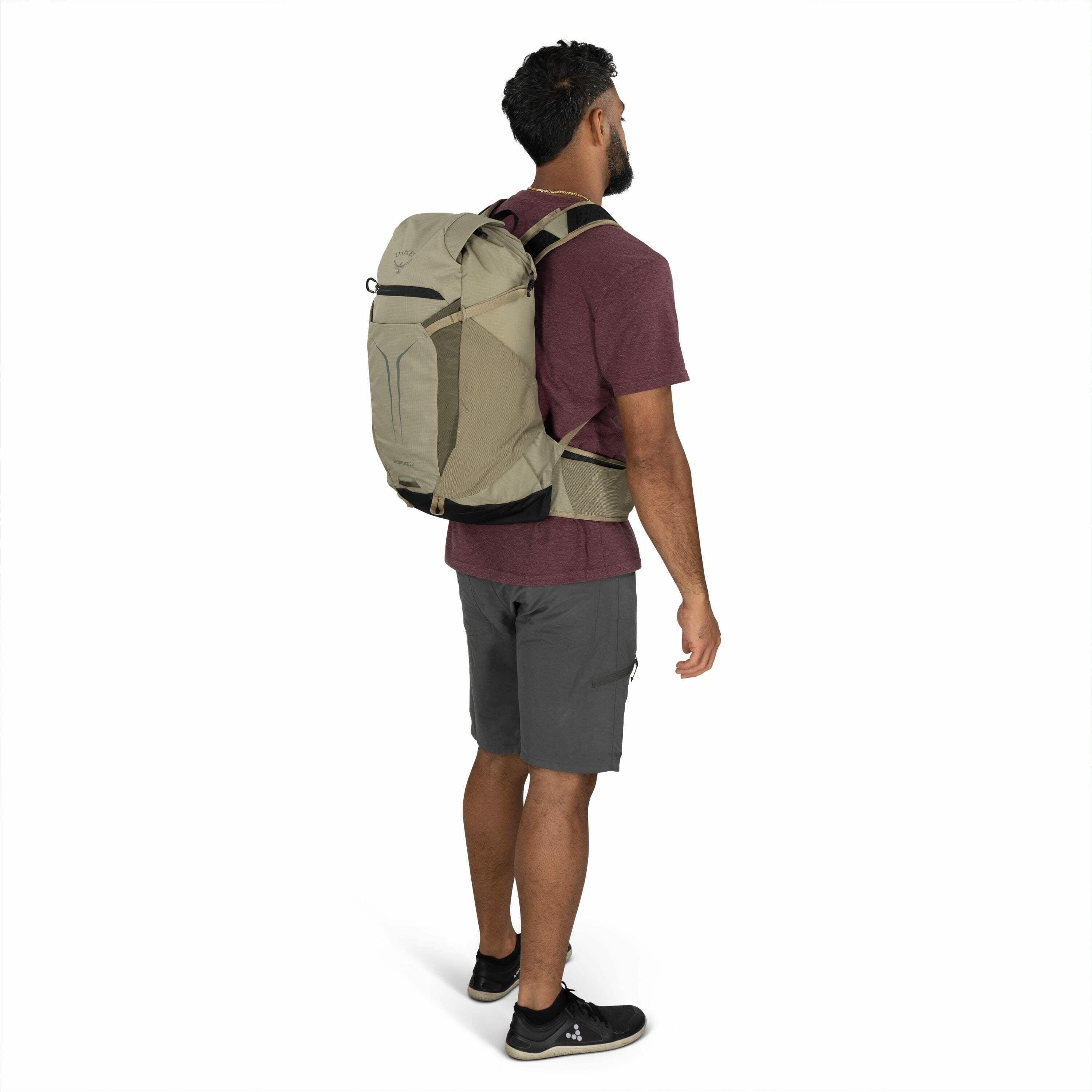 The image shows a man with a short black beard and curly hair standing in a casual pose, wearing a maroon t-shirt and gray shorts. He is equipped with a large, light brown backpack that has a black bottom and visible brand logo, suggesting a design for hi