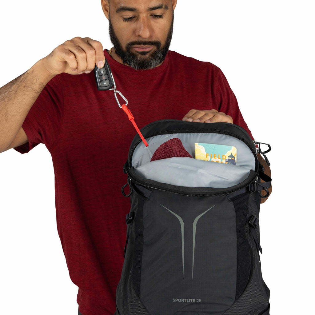 The image features a person wearing a red shirt, holding open a black backpack with "SPORTLITE 25" branding on it. The backpack is partially open, revealing its contents, which include a red knit item, likely a hat or scarf, and a yellow and blue booklet